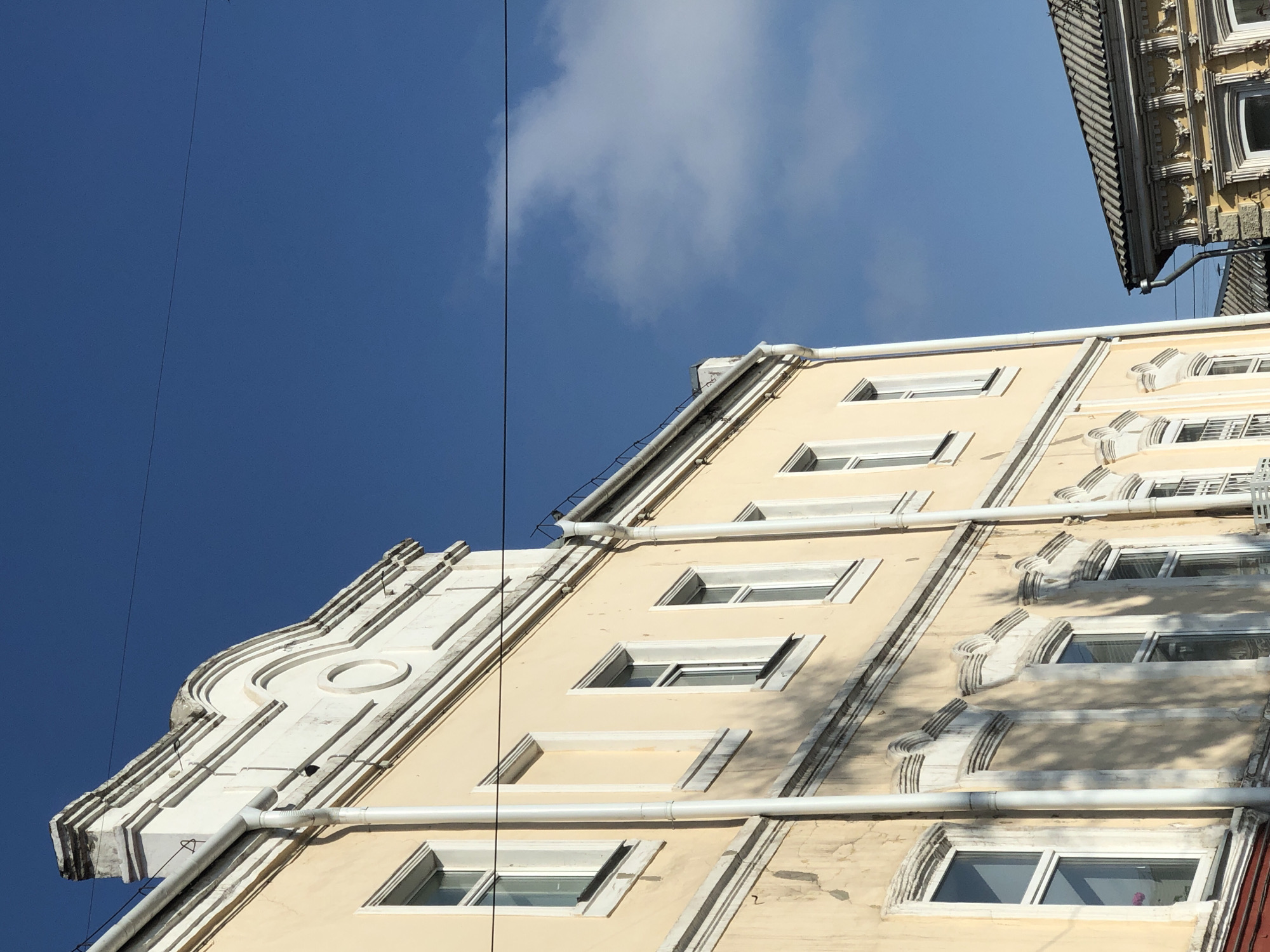 A low-angle shot captures the façade of a light-yellow building with rows of rectangular windows against a clear blue sky with a few scattered clouds. White architectural details frame the windows and run vertically along the building's edges. A white drainpipe runs horizontally across the facade. Electrical wires are visible against the sky. The building appears to be in Krasnoyarsk, Russia.