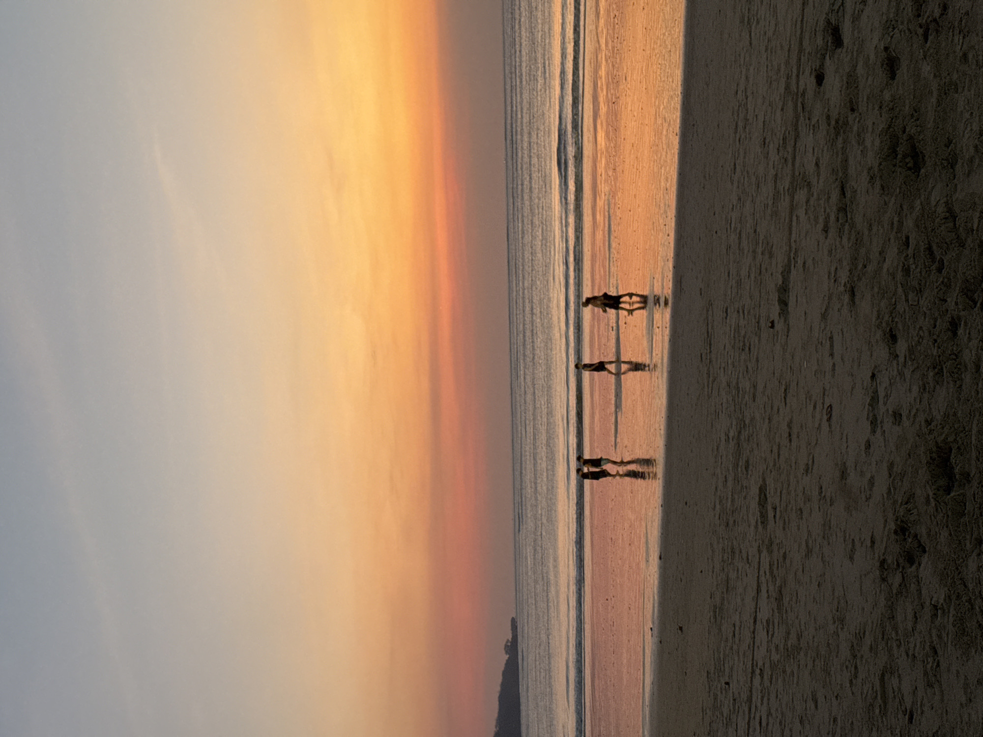 The image depicts a serene beach scene in Kuta Selatan, Indonesia, during sunset. The sky displays a gradient of soft blues and oranges, transitioning into a warm, vibrant hue near the horizon. The ocean is calm, with gentle waves lapping at the shore, reflecting the colorful sky. On the wet sand, a group of four individuals is walking. Two are closer together, appearing to be in conversation, while the other two are further apart, strolling individually. Their silhouettes are cast against the glistening sand. The foreground is dominated by the textured sand of the beach, with the ocean and sky occupying the upper two-thirds of the frame. A distant, dark landmass is visible on the left horizon, adding depth to the scene. The overall impression is one of tranquility and natural beauty at the close of day.