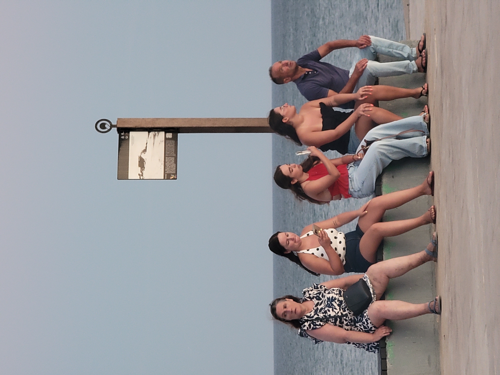 Five people are seated on a low wall overlooking the ocean. From left to right, a woman with a floral dress and dark purse sits with her legs spread. Next to her, a woman in a white polka dot top and denim shorts looks at her phone. To her right, a woman in a red top and light blue jeans holds a phone. A woman in a black strapless top and denim shorts sits next to her. Finally, a man in a blue polo shirt and light blue jeans sits to the far right, looking up and to the right. Behind them, a signpost with a circular emblem and a diagram is visible against a pale sky and the ocean. This scene is likely located on the Quarteira boardwalk in Portugal, given the characteristic coastal setting and urban design elements.