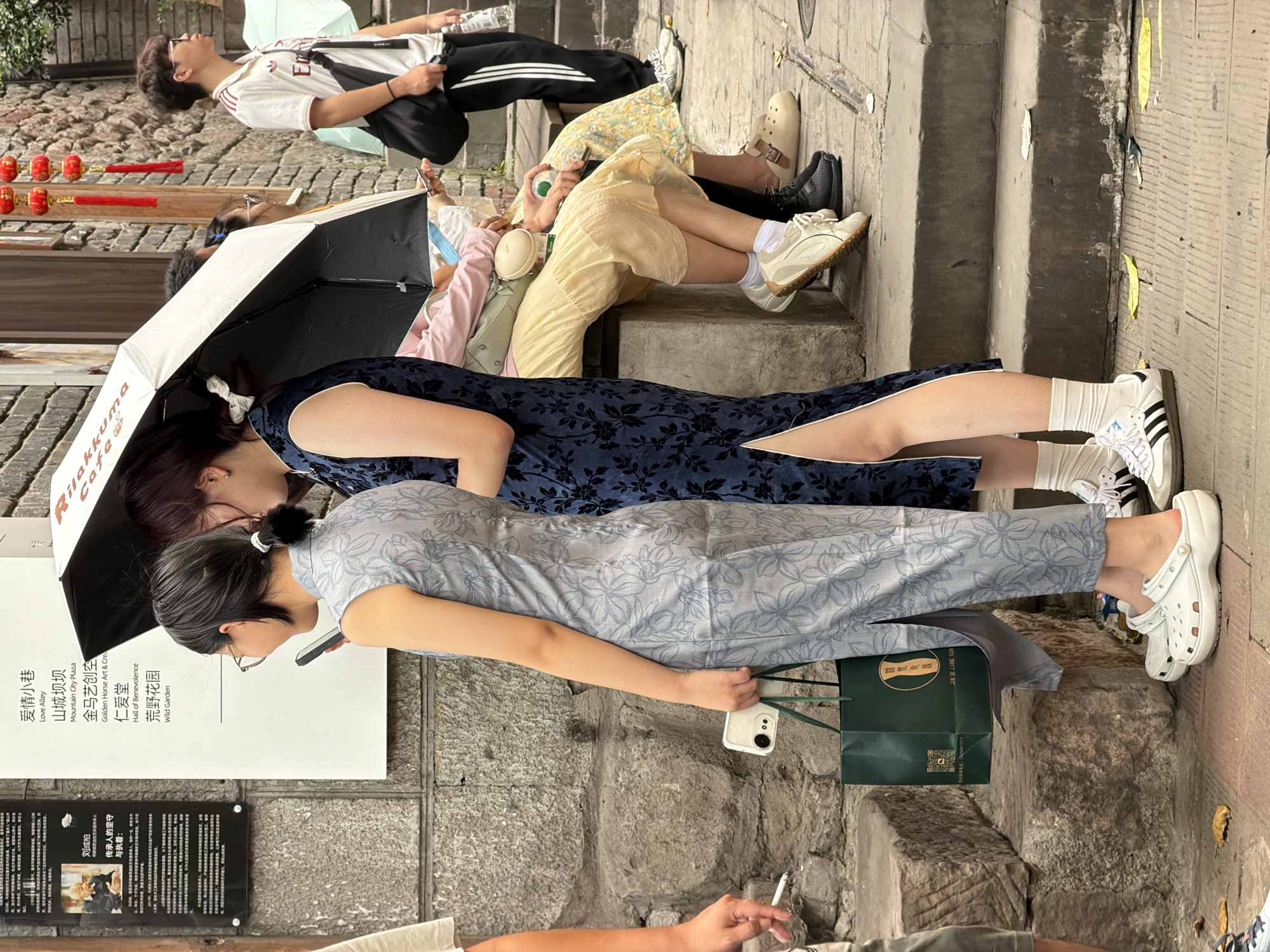 The image captures a street scene in Yuzhong District, China, likely in a historical or cultural area given the traditional architecture and signage. Two young women are the central figures, both dressed in modern interpretations of traditional Chinese qipao dresses. The woman on the left wears a light blue qipao with a floral pattern and glasses, holding a smartphone and a green shopping bag. The woman on the right is in a dark blue qipao with a similar floral motif, holding a black umbrella. They are standing on stone steps, with other individuals visible in the background, including a young man in athletic wear. The environment features stone walls, steps, and potted plants, suggesting an outdoor commercial or tourist area. The overall atmosphere is casual and daylit.