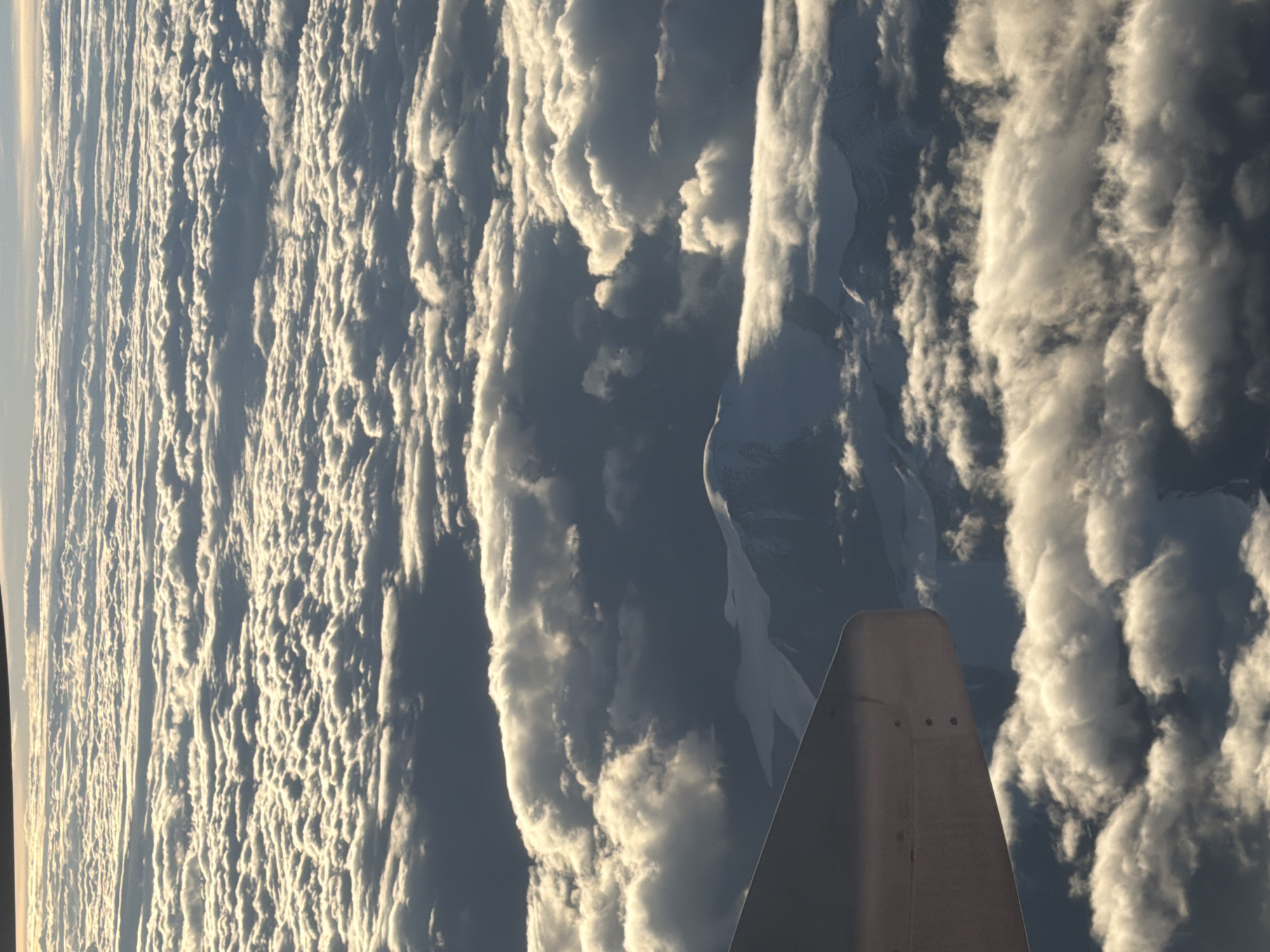An aerial view captures a vast expanse of clouds with sunlight glinting off their surfaces, creating a textured and layered appearance. Partially visible in the foreground is a portion of an aircraft wing, its metallic surface showing signs of wear and fastened with visible rivets. Below the clouds, jagged mountain peaks, capped with snow, emerge from the shadowed landscape. The overall scene is one of natural grandeur, viewed from an elevated perspective, suggesting travel or exploration over a mountainous terrain.