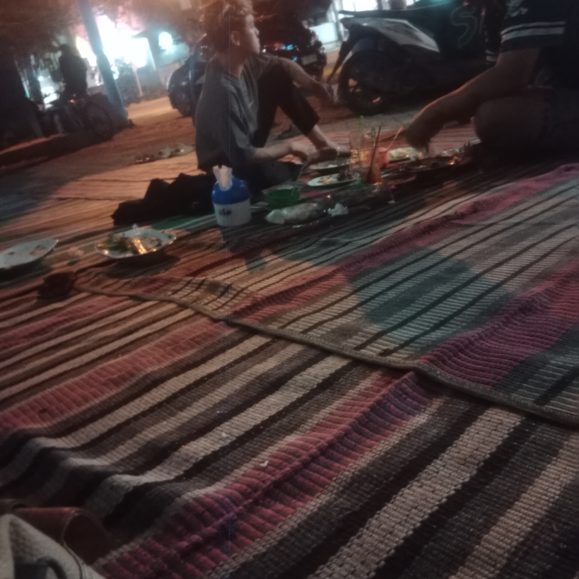 In the dimly lit environment of Pilang Kidul, Indonesia, two individuals are seated on a striped mat on the ground. One person, a male with light-colored hair, is wearing a grey shirt and dark pants, looking off to the side. The other individual, whose body is mostly obscured, is reaching for food laid out on several plates on the mat. The scene suggests a casual outdoor gathering with food and drinks, possibly near a road or street given the presence of a parked motorcycle in the background. The lighting is artificial and casts a warm glow on the scene.