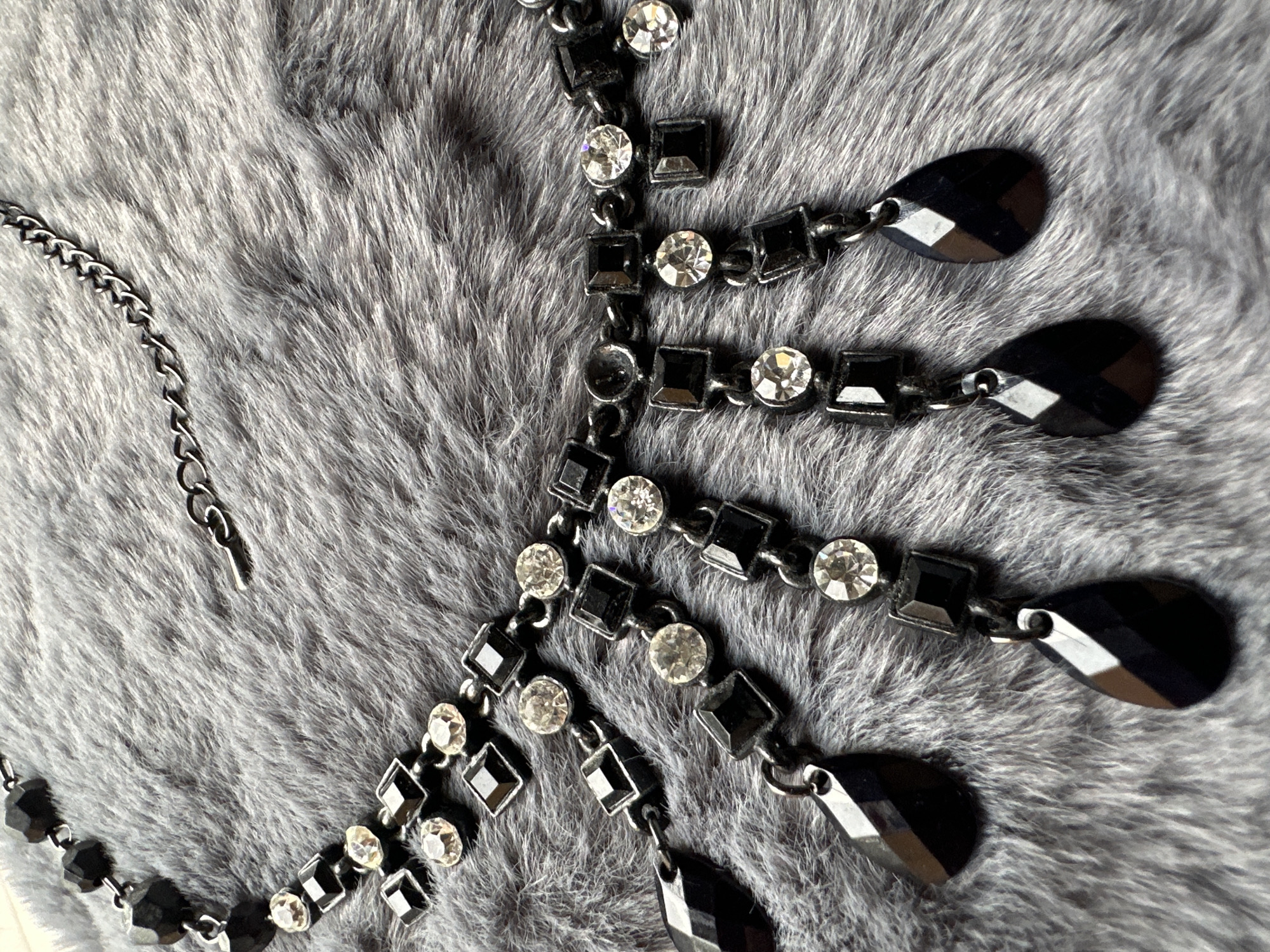 A close-up, overhead shot displays a black beaded necklace with clear rhinestones and black gemstone pendants, resting on a soft gray furry surface. The necklace is ornate, featuring a chain with various geometric charms, including faceted black square beads, clear circular rhinestones, and black teardrop-shaped pendants. The furry background appears to be a throw or rug. The location is not specified in the city of Unknown, Unknown.