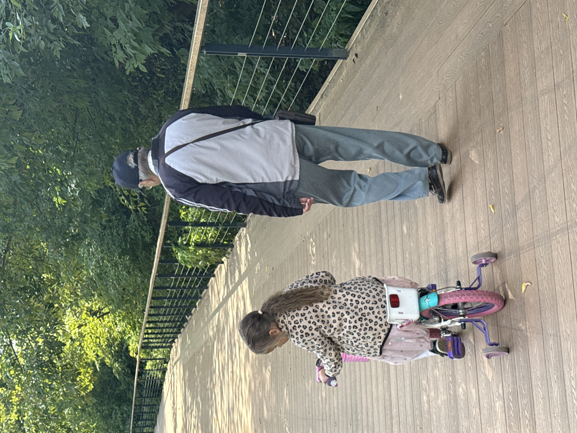 An overhead shot captures an elderly man and a young girl on a wooden boardwalk that curves to the left. The man, viewed from behind, wears a dark baseball cap, a jacket with gray and black panels, and light blue pants. He has a dark bag slung across his right shoulder. The girl, also viewed from behind, sits on a small pink and blue bicycle with training wheels. She is wearing a leopard-print jacket over a light pink skirt. Her dark hair is pulled back into a ponytail. The boardwalk is bordered by a black metal railing on the left and dense green foliage on the right. The scene is illuminated by sunlight, casting dappled shadows on the wooden planks. This image does not provide enough information to determine its location in Moscow, Russia.