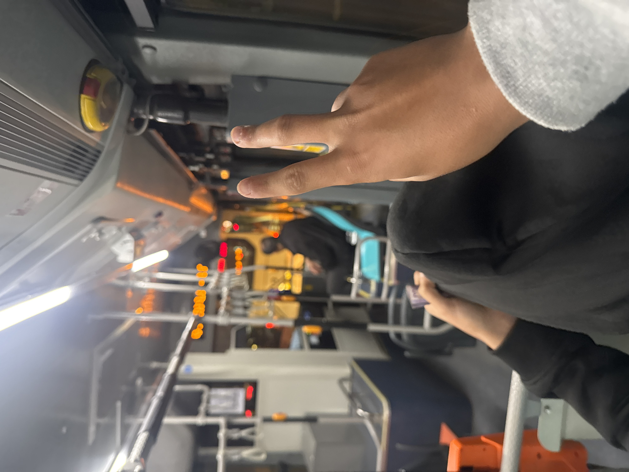 The image shows a person sitting on a bus. The bus is traveling through Yerevan, Armenia at night. The person is wearing a gray sweatshirt and black pants. They are holding up two fingers. Other passengers are visible in the background. The bus interior is lit with artificial light.