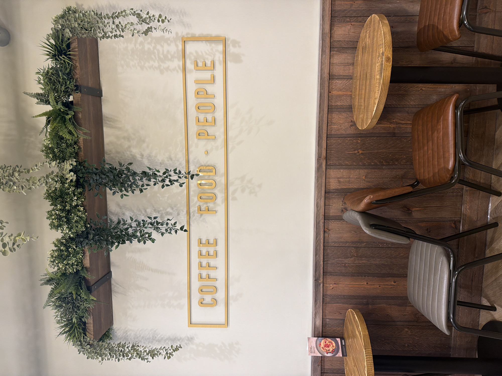 The image shows a section of a cafe interior. The left side of the frame features a white wall with a vertical wooden beam adorned with various green plants. To the right of this arrangement is a gold-framed sign with the words "COFFEE - FOOD - PEOPLE" written vertically. The right side of the image is dominated by a wood-paneled wall with several stools and chairs arranged around it. One stool has a light brown wooden seat, while the chairs have brown leather seats with black metal frames. A menu card is also visible near the bottom right corner of the image. The cafe's specific location in Mole Valley, United Kingdom cannot be determined from the image.