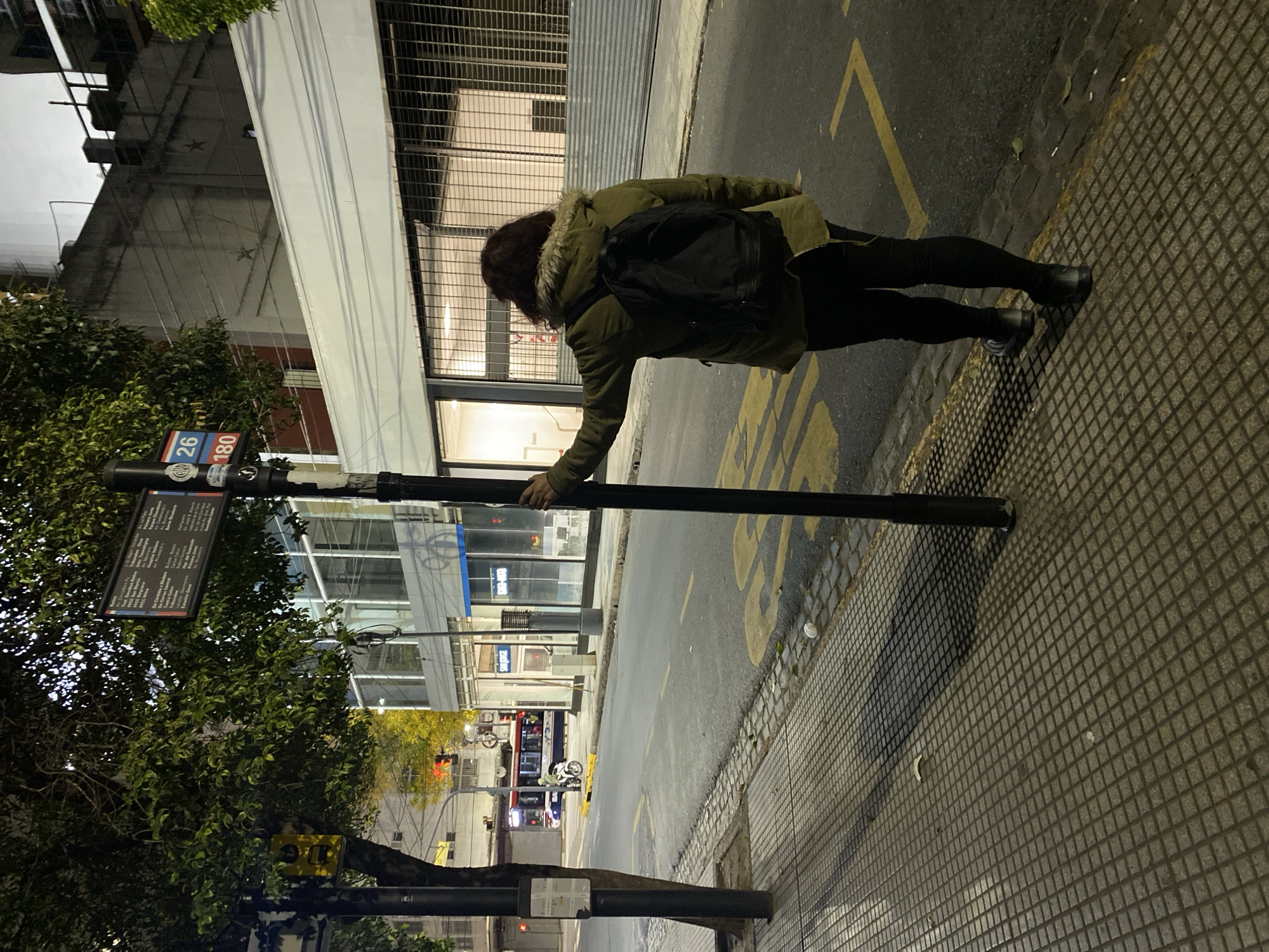 A woman wearing a green jacket and black backpack is standing on a patterned sidewalk next to a street in Buenos Aires, Argentina. She is touching a black pole with a bus schedule sign. A tree is next to the pole. The street has yellow lane markings and a curb. A bus and motorcycle are visible in the distance. Buildings line the street. One building has metal shutters and another has a blue sign. Bus routes 26 and 180 are visible on the bus stop sign.