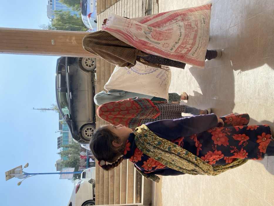 The image depicts an outdoor urban scene under bright daylight in Kandahar, Afghanistan. In the immediate foreground, a young girl, approximately 5-8 years old, is shown in right profile, her dark hair styled in two braids with a white, heart-shaped hair tie visible on the braid closer to the viewer. She wears a dark blue long-sleeved garment adorned with a vibrant red floral pattern and a green, yellow, and brown patterned scarf draped over her left shoulder.
Behind her, a line of at least four additional individuals, mostly obscured from the waist up by head coverings, proceed from left to right. The person directly behind the foreground girl wears a red patterned head covering. The next individual wears a light blue head covering. The fourth person in line wears a brown patterned head covering and carries a large white sack over their left shoulder, marked with partial text including "0706". The final visible person in the queue, positioned to the far right, carries a large white and red patterned sack over their right shoulder. This sack prominently displays text in Dari/Pashto, indicating "٥٠ كيلوجرام" (50 kilograms) and "افغان" (Afghan), suggesting it contains bulk goods like flour.
The ground is a light-colored, paved surface, reflecting strong shadows from the individuals. In the mid-ground, a series of light brown concrete steps ascend from left to right. Above and behind these steps, a black hatchback car, partially dusty, and a white sedan are visible on a raised roadway. To the right, a vertical light brown concrete pillar stands adjacent to an ascending concrete ramp. The background includes a tall blue lamppost with solar panels, distant buildings with visible solar installations, scattered trees, and a cellular tower against a clear blue sky. The specific street or precise location within Kandahar is not identifiable from the visual evidence.