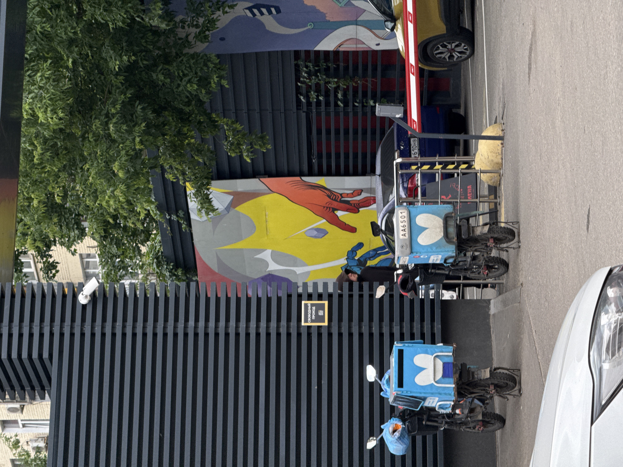 The image depicts an outdoor scene in Moscow, Russia, likely near a business or residential complex. In the foreground, a white car's hood and headlight are visible. Behind it, two blue delivery scooters with insulated cargo boxes are parked. The scooters are branded with a white heart logo. A person in a dark hooded jacket stands near one of the scooters, partially obscured by a metal railing.
The background features a modern building with dark horizontal siding, contrasting with a vibrant mural on the wall. The mural is abstract, with bold colors like yellow, red, blue, and purple, and features a large red hand and geometric shapes. A white security camera is mounted on the building's siding. A red and white striped barrier arm is lowered, indicating a checkpoint or entry. Behind the barrier, a dark blue car and a yellow car are visible, suggesting a parking area. Trees with green foliage overhang the scene. A small sign with Cyrillic text, reading "СВОБОДНЫЙ ПРОХОД" (Free Passage), is attached to the dark siding. The overall environment appears to be a functional urban space, possibly a service entrance or loading zone.