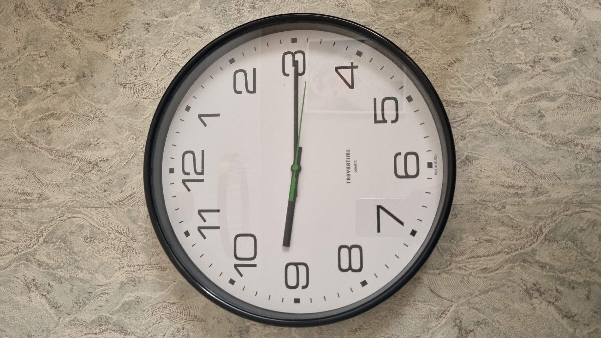 The image shows a round analog clock with a black frame hanging on a textured wallpaper. The clock face is white with black Arabic numerals. The hour hand is pointing to the 9, and the minute hand is pointing to the 3. The second hand is green. The brand name "TAOYHATIME QUARTZ" and "MADE IN BELARUS" are visible at the bottom of the clock face. The background wallpaper has a marbled pattern in shades of gray and beige. There are no people or animals present in the image. The lighting suggests an indoor setting, possibly a room in a house or office. The clock is positioned centrally in the frame.