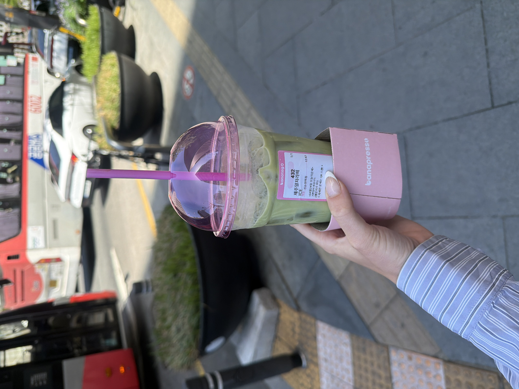 A person with a striped light blue long-sleeved shirt holds a pink-lidded cup with a pink straw filled with a green drink and a pink cardboard cup holder with the word "banapress" on it. The drink label states: "banatag" "432" "1/1" "제주말차라떼" "ICE" "매장 신논현역점" "<04월24일 오늘의운세>" "기쁜일이 생깁니다" "(행운지수 85)" "*자세한 운세는 QR 스캔!*" "12:47". In the background, a bus and white car are visible. There are black planters with greenery near the bus. The location is in the city of Seoul, South Korea.