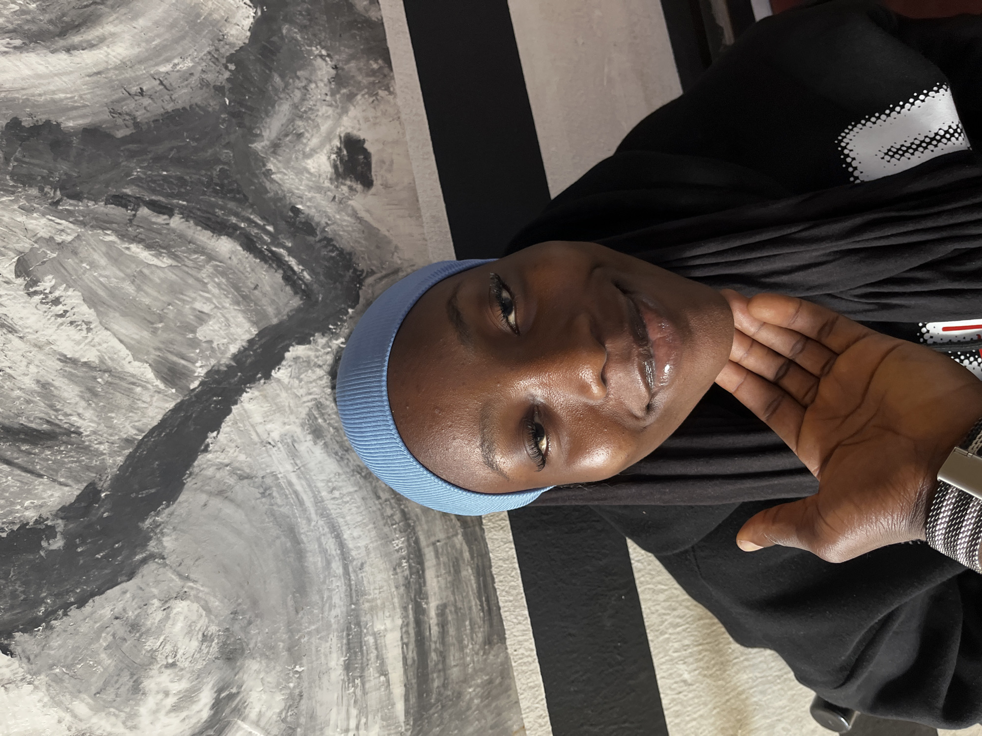A young woman with dark skin is captured in a close-up, head-and-shoulders portrait, looking directly at the viewer with a faint, pleasant smile. She is wearing a light blue, close-fitting head covering, possibly a beanie or underscarf, and a black long-sleeved top or hoodie. A white, abstract dotted pattern is visible on the sleeve of her black top. Her left hand, adorned with a silver-toned mesh strap watch, is raised near her face, with fingers slightly parted. Her skin shows a natural sheen, and her lips appear glossy.
The background features an abstract wall design. On the left side of the frame, the wall is painted with broad, swirling brushstrokes of various shades of grey and white, creating a textured appearance. To the right of this section, behind the woman, the wall transitions into a pattern of alternating vertical black and white stripes. The setting appears to be indoors. No discernible text is visible on her clothing or in the background. The image evokes a calm and friendly mood. The location context for this image is Jos, Nigeria.