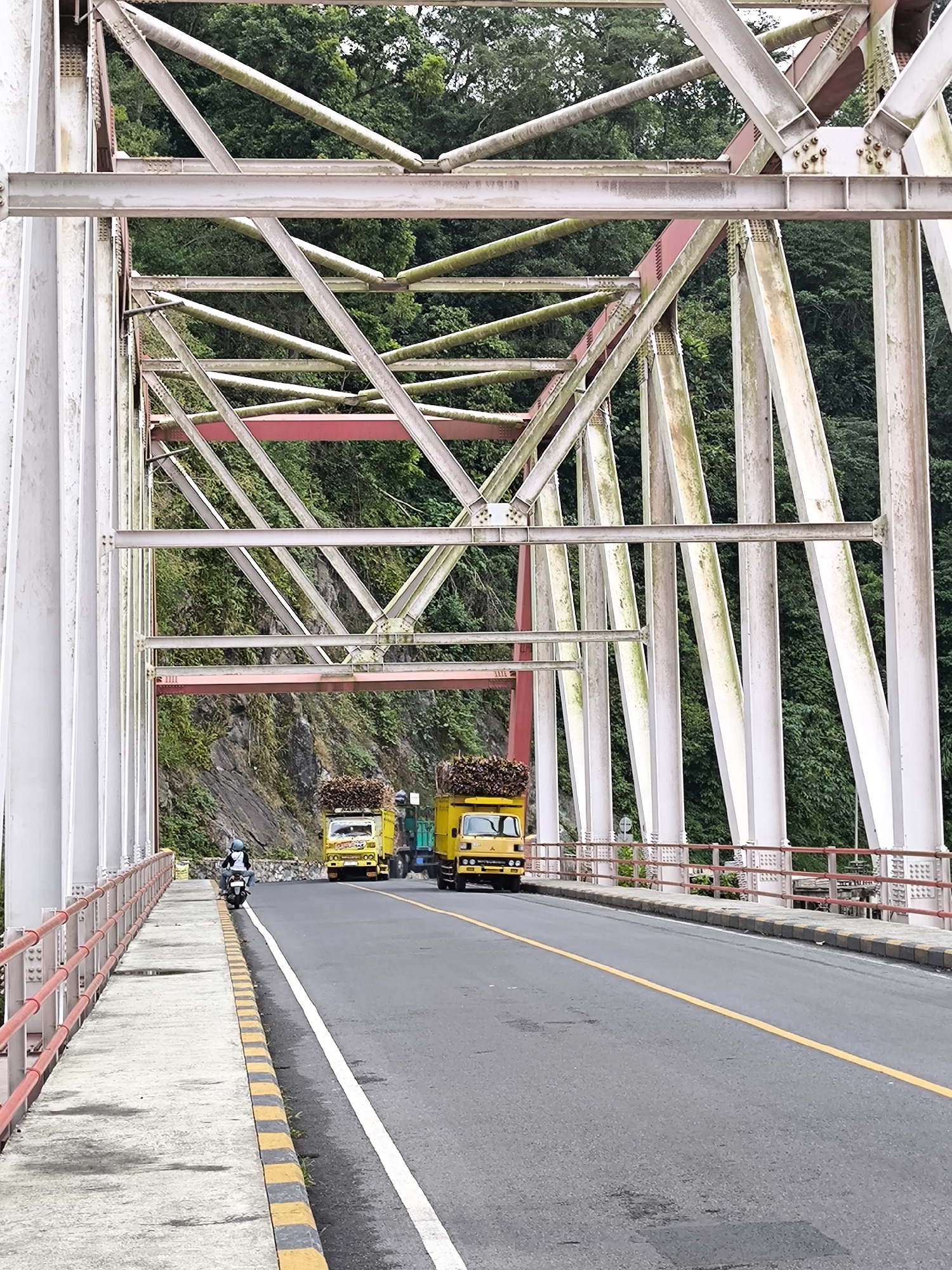 The image shows a bridge in Supiturang, Indonesia, with two yellow trucks carrying bundles of wood on their beds, driving on the road. A person is riding a motorcycle on the pedestrian walkway of the bridge. The bridge has a metal truss structure painted in white and red, and it spans across a forested area. The road is asphalt with a yellow line dividing the lanes. The sky is overcast.