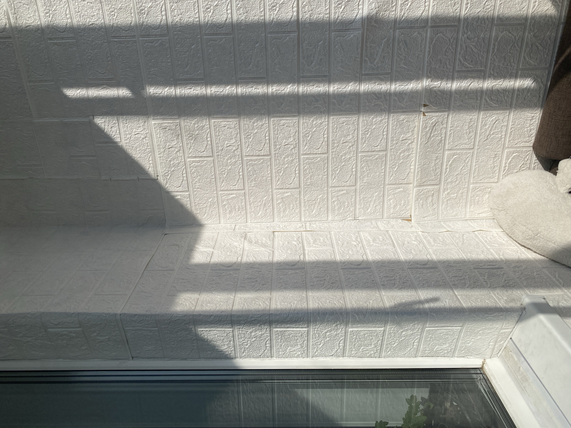 The image depicts a corner of an interior space with a white brick-patterned wallpaper. Sunlight streams in from the left, casting distinct shadows of window blinds onto the wall. A window with a dark frame is visible on the left side of the image, and a white windowsill runs along the bottom. The wallpaper shows some minor imperfections, with a few small areas where the surface appears to be peeling or damaged. In the bottom right corner, a fluffy white object, possibly a stuffed animal, is partially visible resting on a brown surface, likely furniture. The scene is clean and uncluttered, suggesting a domestic or commercial interior. The specific location in Novosibirsk, Russia, cannot be determined from the visual information provided in the image.