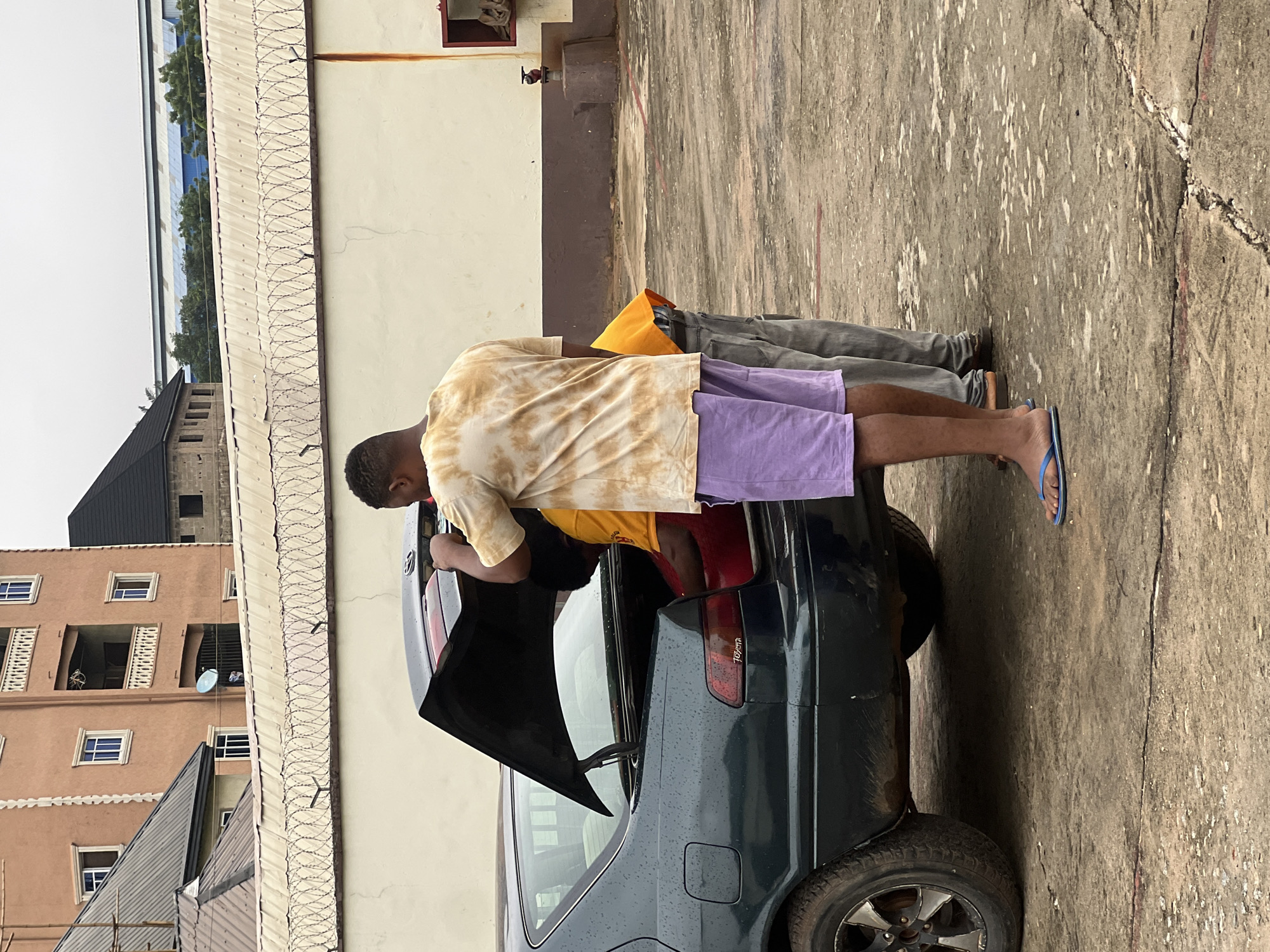 In Nkpor, Nigeria, two individuals are seen in a lot adjacent to a building. The primary focus is a dark-colored sedan with its trunk open. One person, wearing a tie-dye t-shirt and purple shorts, stands beside the trunk, appearing to be interacting with its contents. Another person, wearing a yellow t-shirt and grey trousers, is partially visible inside the trunk, also seemingly engaged with the items within. The ground is paved with concrete, exhibiting some wear and tear. In the background, a multi-story building with orange-colored walls and balconies stands, along with a barbed wire fence, indicative of security measures. The sky is overcast, suggesting a cloudy day. The overall scene depicts a moment of loading or unloading of items from the car.