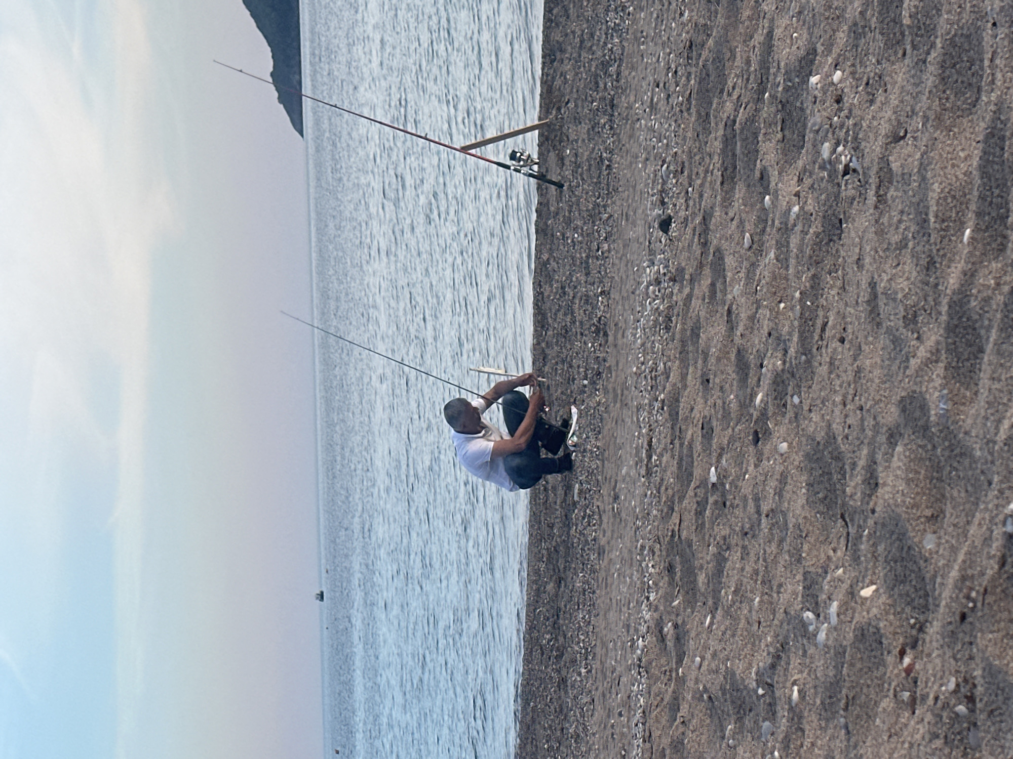 A man wearing a white shirt and dark jeans sits on a beach in Budva, Montenegro, next to the water with his fishing rod. Another fishing rod is standing in the sand farther down the beach. The beach is mostly dark sand and small pebbles. The water is blue and there is a mountainous landmass in the distance. The sky is light blue to white with a light haze.