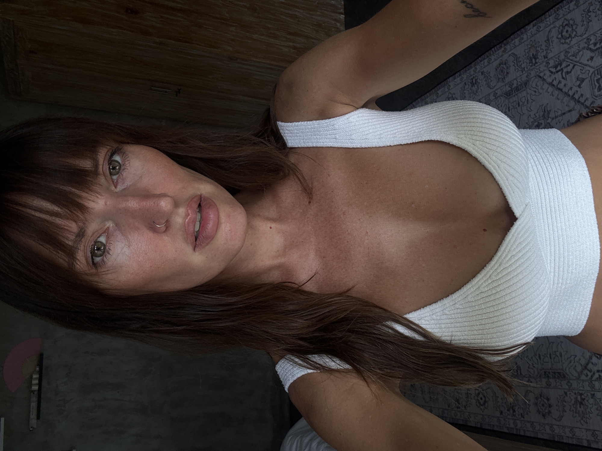 A young woman is centered in a low-angle, close-up selfie indoors. She wears a white, ribbed, cropped tank top. She has fair skin, brown hair with bangs, and a nose ring. Her hair is partially covering the left side of her face. A portion of a tattoo is visible on her right shoulder. Behind her is a gray rug. To the left, a gray wall with a pink fan is visible. Above and to the left is a wooden surface. The location is potentially within a residential or indoor setting in Tibubeneng, Indonesia.