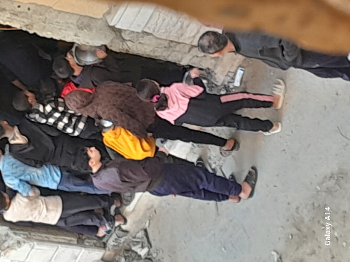 The image depicts a gathering of approximately ten individuals, predominantly males and children, in a damaged urban environment within Khan Yunis, Palestinian Territory. The ground consists of concrete or asphalt scattered with debris, including small rocks and fragments of building material. A broken concrete wall or structure is visible on the right side and upper left. In the background, multiple individuals are leaning into or looking intently into a dark, narrow recess or void between structures. Attire among these individuals includes a light-colored long-sleeved shirt, a light blue long-sleeved shirt, a black hooded sweatshirt, and a black and white checkered shirt. In the middle ground, a male in a dark purple top and dark pants stands, looking towards the recessed area. To his right, an individual wearing a brown patterned hooded jacket and black pants holds a yellow item, possibly a garment or bag, and directs their gaze towards the same dark opening. A young female, viewed from behind, wears a black tracksuit with pink stripes on the pants and stands facing left. A white cylindrical object lies on the ground near her feet. On the far right, a male in dark clothing is partially visible, also observing the scene. The collective posture and attention of the individuals are focused on the dark, confined space.