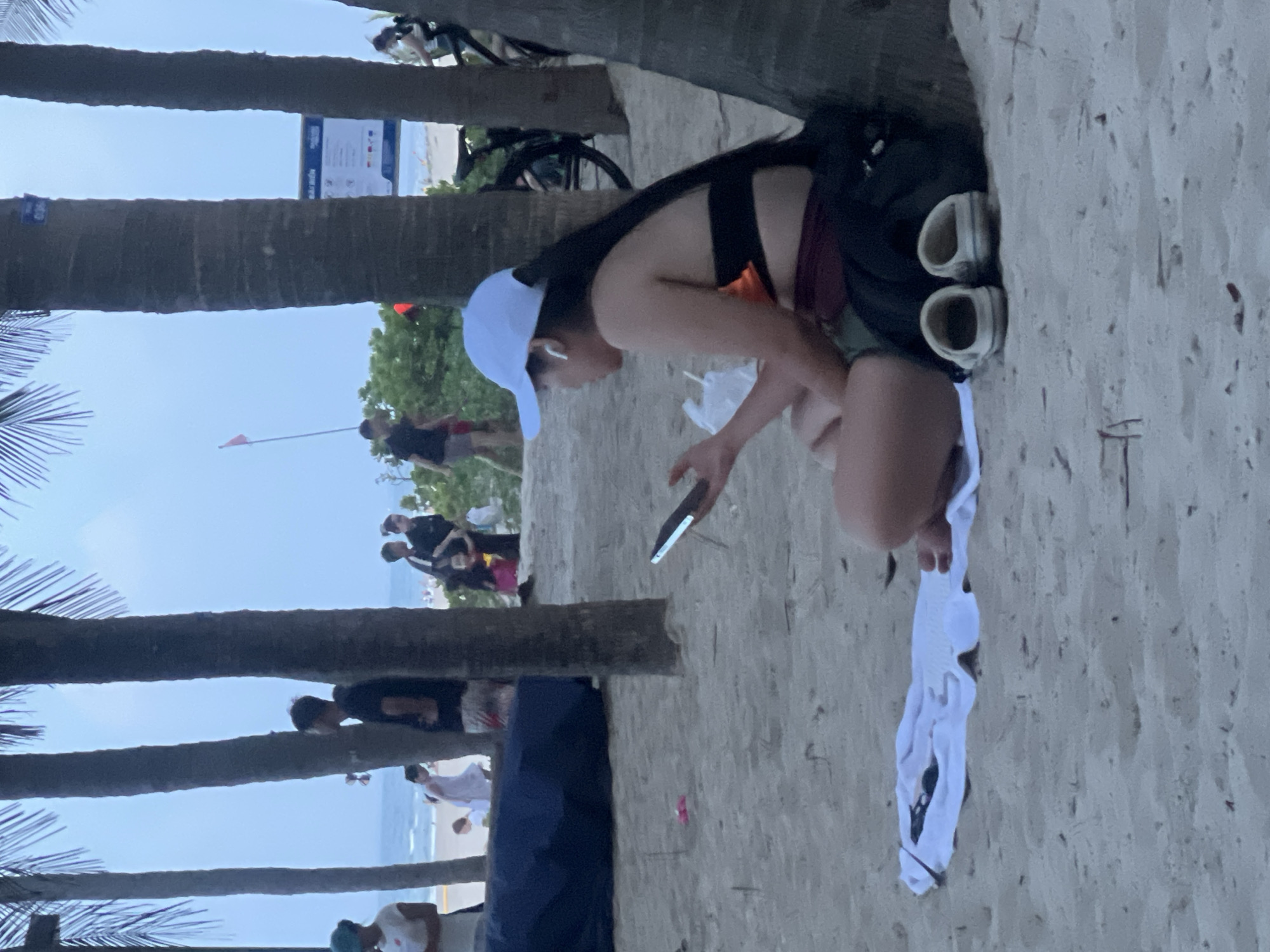 On a sandy beach, likely at My Khe Beach in Da Nang, Vietnam, a young woman sits partially leaning against a palm tree. She is wearing a white baseball cap, an orange bikini top with a black band, and burgundy bottoms. She has long black hair and is using wireless earbuds while looking at her phone. Beside her are two white clogs and a white towel. In the background, other beachgoers are visible, as well as palm trees, greenery, and the ocean under an overcast sky. A bicycle is also visible to the right.