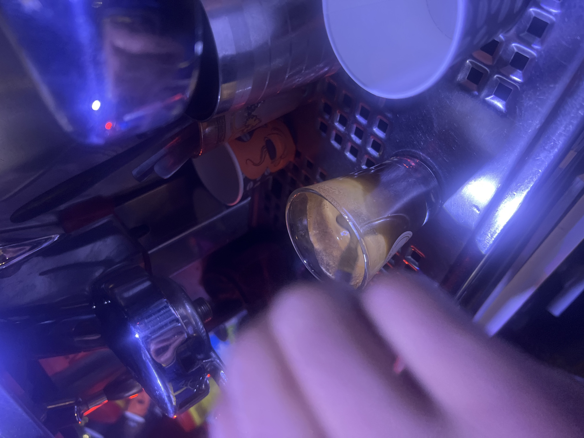 This close-up, low-angle shot depicts a beverage preparation area within an indoor commercial setting, likely a cafe or bar. The scene is characterized by highly reflective metallic surfaces, possibly belonging to an espresso machine or a drink dispenser, bathed in strong blue and purple artificial lighting.
Various objects are visible: A white paper cup is positioned at the top right. Beneath it, an orange cup with a black, stylized face or character design is partially discernible. Centrally to the right, a clear glass holds a yellowish-brown liquid, consistent with coffee or a mixed drink, exhibiting some foam or bubbles. On the bottom left, a chrome-plated spout or dispensing head from a machine gleams. Bright white and red indicator lights are visible as reflections on the metallic surfaces.
A blurred human hand extends into the foreground from the bottom right, suggesting an active interaction with the machine or the drink. The vibrant, artificial lighting creates a modern and possibly evening or nighttime atmosphere. No explicit text is visible. The location context is Martil, Morocco.