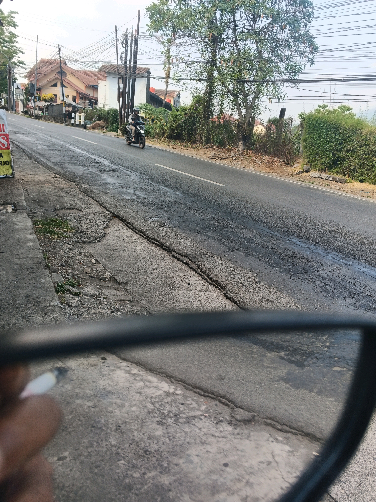 The image depicts a street scene in Ungaran, Indonesia. In the foreground, a person's hand is holding a lit cigarette, partially visible within a rearview mirror's frame. The mirror reflects a paved road with visible cracks and patches. A motorcycle with a rider wearing a helmet is traveling away from the viewer on the road. On the left side of the road, there is a sidewalk with some weeds and debris. Beyond the sidewalk, a building with a tiled roof is visible, along with trees and utility poles with numerous overhead wires. The environment appears to be a semi-urban area with residential structures and infrastructure. The lighting suggests it is daytime.