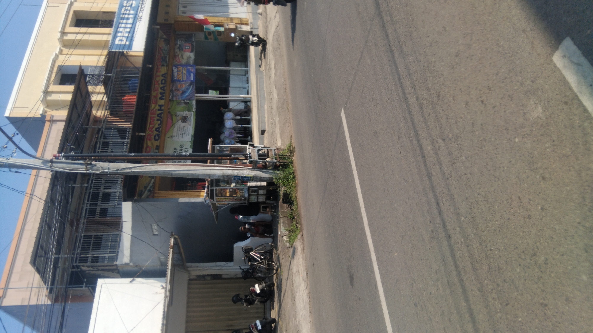 The image shows a street scene in Proyonanggan Tengah, Indonesia. In the foreground is a paved road with a white line marking. To the left of the road is a sidewalk with several parked motorcycles and a bicycle. Further back, there is a building with multiple stories. The ground floor of the building appears to be a store or business with merchandise displayed in the windows. People can be seen inside the store and near a small kiosk on the sidewalk. A utility pole with wires runs vertically through the image. The sky is clear and blue, indicating it is daytime. The overall scene depicts a typical urban environment with commercial activity and transportation.