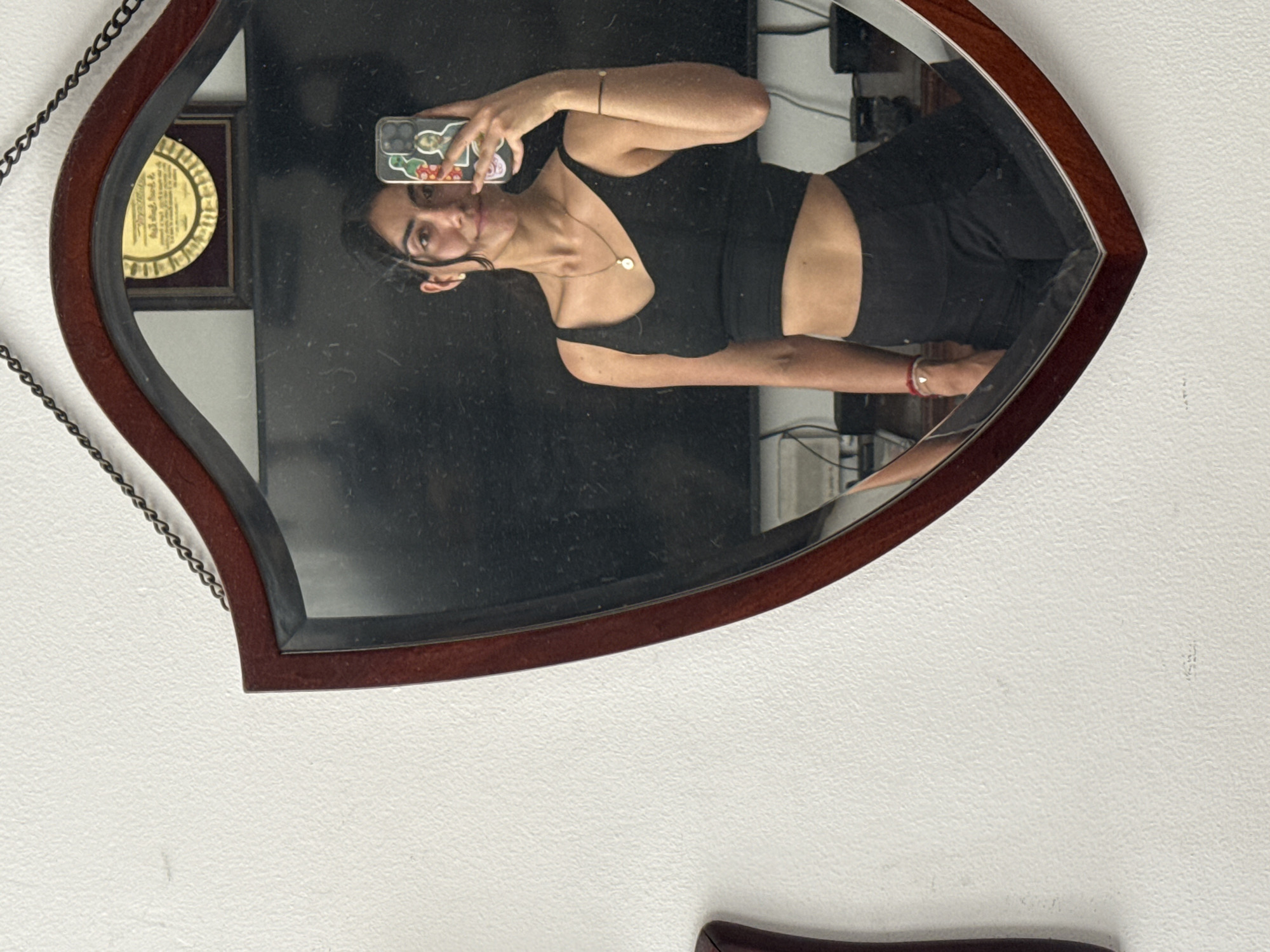 The image, rotated 90 degrees counter-clockwise, captures a mirror selfie taken by a person in an indoor setting in Puebla, Mexico. The subject, likely female, has dark hair and is wearing a black sleeveless top and black bottoms. She is accessorized with a necklace featuring a round pendant and bracelets on both wrists. She holds a smartphone, encased in a colorful cover adorned with cartoonish figures, to take the photo. Her expression is neutral or slightly smiling.
The mirror itself has a distinctive eye or leaf-like shape, framed in dark brown wood with a narrow silver accent. A dark metal chain hangs from the upper left side of the frame. The mirror surface appears somewhat dusty or smudged.
In the reflection, a gold-colored framed award or plaque with illegible text is visible on a wall to the left of the person's head. Further back in the reflection, some electronic devices and cables can be seen, suggesting a room with media or office equipment. The wall visible outside the mirror's frame is white and has a textured finish. The scene portrays a casual moment in what appears to be a home or apartment.