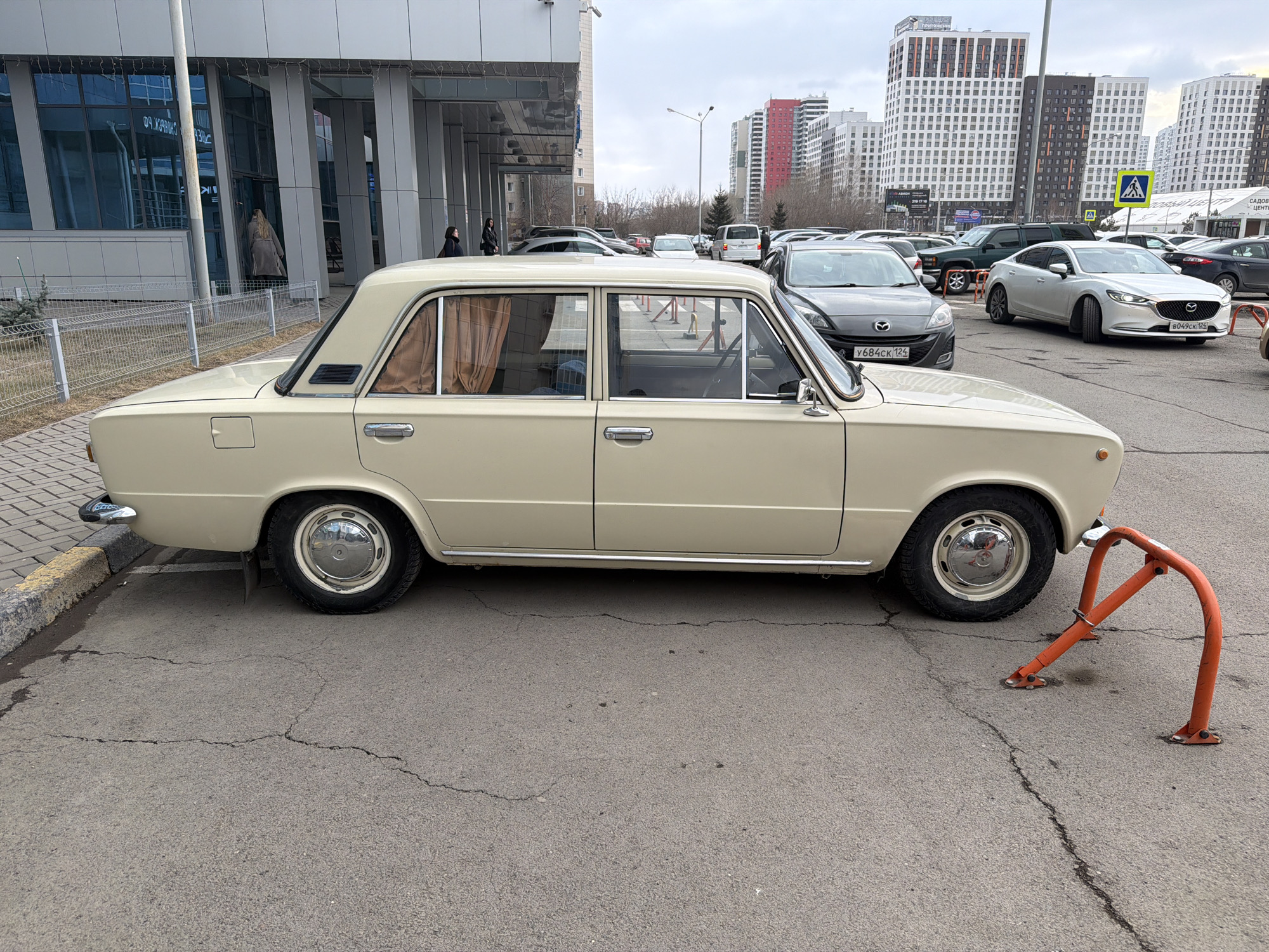 The image shows a beige VAZ-2106 parked on cracked asphalt in what appears to be a parking lot in Krasnoyarsk, Russia. The car is positioned parallel to the curb and faces right. An orange metal parking barrier rests against its front tire. The license plate of a gray Mazda is partially visible in the background. Behind the Mazda, several other cars are parked. Modern, multi-story residential buildings dominate the background. The building next to the car is a modern structure with a glass facade. People are seen entering and exiting the building. The sky is overcast.