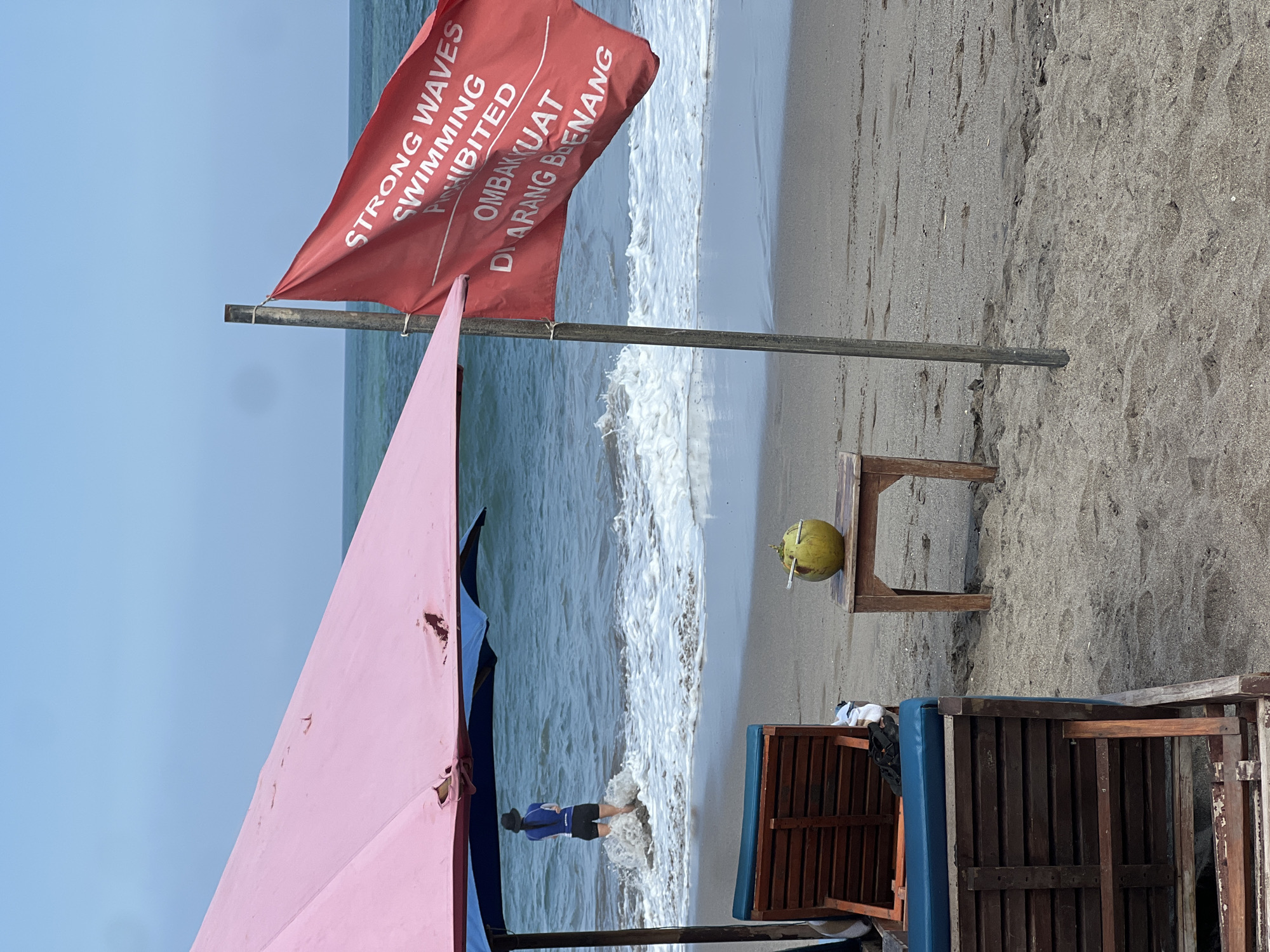 A person stands in the ocean waves on a beach in North Kuta, Indonesia. The person is wearing a dark hat, a blue long-sleeved shirt, and black shorts. A large pink umbrella, possibly torn in places, is in the foreground, casting a shadow over two wooden lounge chairs. A small wooden table sits on the sand to the right of the chairs, holding a green coconut with a straw inserted. A red flag with white lettering, reading "STRONG WAVES SWIMMING PROHIBITED" and "OMBAK KUAT DI LARANG BERENANG", is attached to a wooden pole to the right of the table. The ocean is choppy with white waves crashing on the shore. The sky is a pale blue. The sand is light brown.