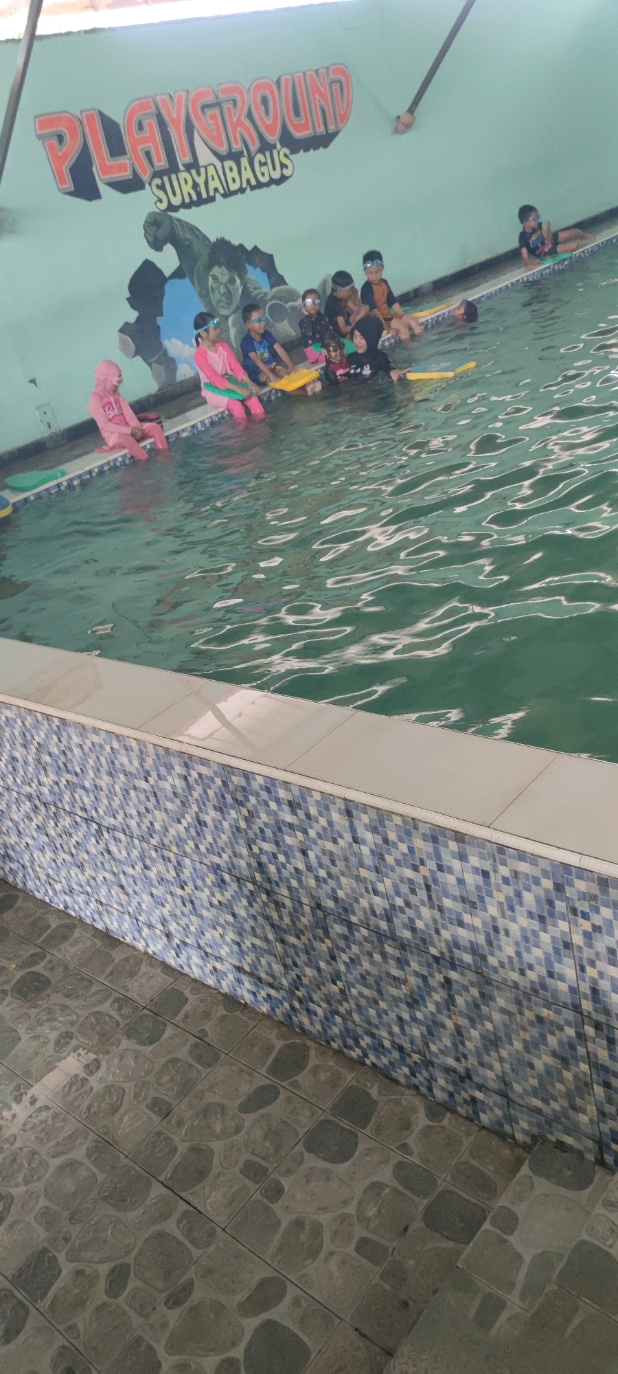 A group of children are gathered in a swimming pool at a playground in Jemundo, Indonesia. The wall behind them is decorated with a mural of a superhero, and the words "PLAYGROUND SURYA BAGUS" are painted in large, colorful letters. The children are wearing swimsuits and goggles, and some are holding yellow flotation devices. The water is green and appears to be shallow. The pool area is tiled with blue and white mosaic tiles. The floor surrounding the pool is paved with stone-like tiles.
