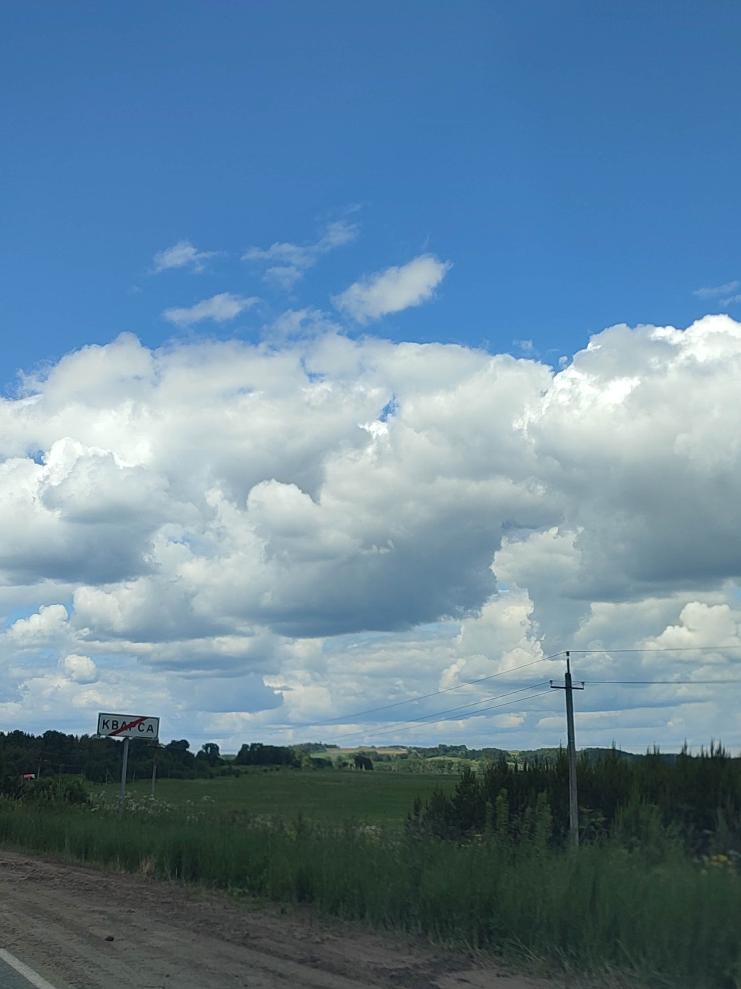 This media file captures a scenic rural landscape under a bright blue sky dotted with fluffy white clouds. The foreground features a dirt road, suggesting an outdoor setting likely in a rural or countryside area. To the left, a road sign with red and white markings is visible, bearing the Cyrillic text "КВАРСА" with a red diagonal line through it, indicating this is likely an entrance to or exit from a settlement or a place with that name.
Beyond the road, a vast expanse of green fields stretches out, interspersed with trees and vegetation. In the distance, rolling hills form the horizon. A solitary utility pole with power lines stands on the right side of the frame, adding to the rural atmosphere. The lighting suggests it is daytime, with the sun illuminating the landscape and casting shadows, though the exact time of day is difficult to pinpoint. The weather appears pleasant, with the clear sky and prominent clouds indicating a fair day. There are no people visible in the image, and the overall mood is peaceful and serene, characteristic of a quiet, natural environment.