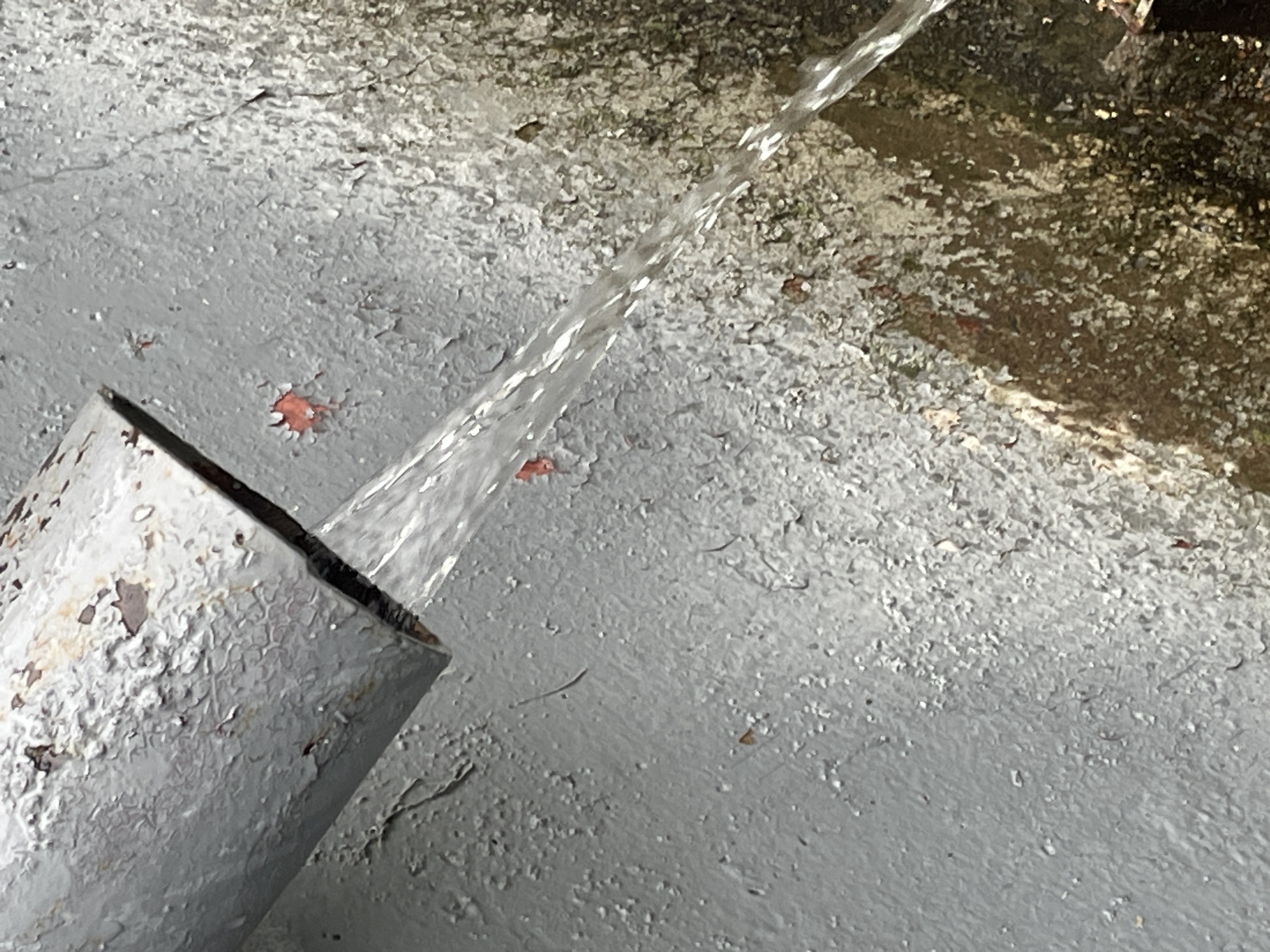 A close-up, high-angle shot shows a stream of clear water flowing from a rusty, gray pipe. The pipe is angled downwards and to the right. The water is breaking into many small droplets as it exits the pipe. The surface below the pipe is a rough, weathered gray wall with peeling paint. Patches of reddish-brown rust are visible beneath the peeling gray paint. The wall appears damp and stained in some areas, with darker, possibly organic material, visible in the lower right section of the image. The scene is likely outdoors and appears to be an external utility pipe. The exact location in Krasnogorsk, Russia cannot be determined from the image alone.
