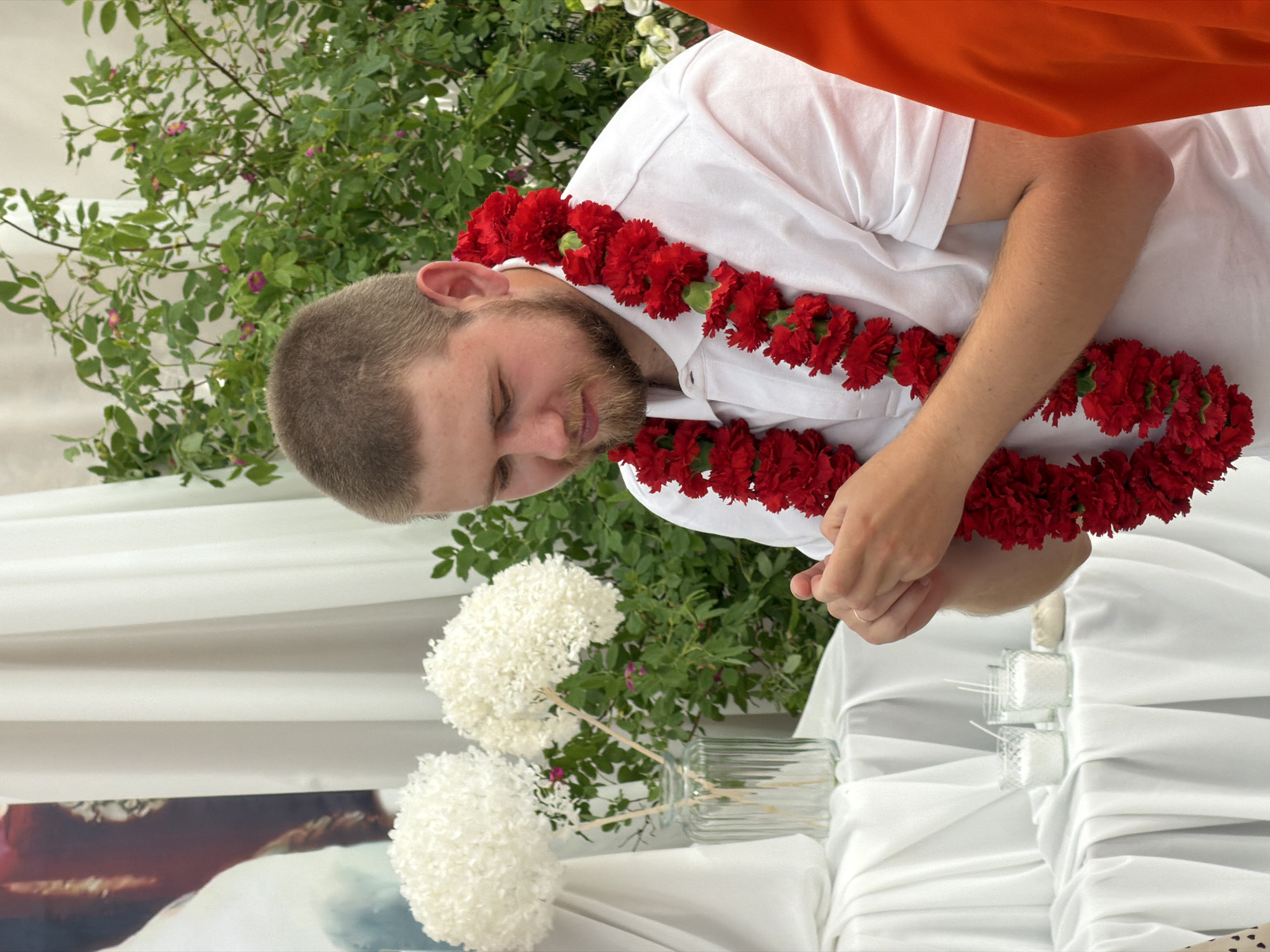 A young Caucasian man with short brown hair and a beard, wearing a white polo shirt adorned with a thick red floral garland, is captured in a medium shot from the chest up. His hands are clasped together in front of him as he looks down with a soft expression. To the left of the man, two white floral arrangements resembling hydrangeas are displayed in a clear ribbed vase. The setting appears to be an outdoor event with lush green foliage and small pink flowers in the background, suggesting a garden or natural setting. A white draped cloth covers a surface in the foreground, on which two small glass containers with white granular substance, possibly salt or sugar, and a few white candles are placed. The man is the central focus, with his attire and the garland suggesting a ceremonial or festive occasion.