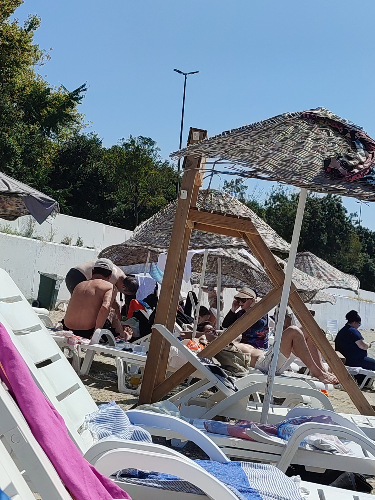 The image depicts a beach scene on a sunny day. Several people are visible, mostly seated or lounging on white plastic beach chairs and under straw-covered umbrellas. In the foreground, white beach chairs are arranged, some with towels draped over them. In the mid-ground, a group of people are gathered around a wooden structure that supports a large umbrella. Some individuals are interacting, with one person appearing to hand something to another. A green trash bin is situated near a white wall in the background. Trees and foliage are visible behind the wall, and the sky is a clear blue. The overall impression is a relaxed atmosphere at a beach resort. This scene could be located at one of the beaches in Kadıköy, Turkey, such as Caddebostan beach or Fenerbahçe beach, which are popular recreational areas in the district.