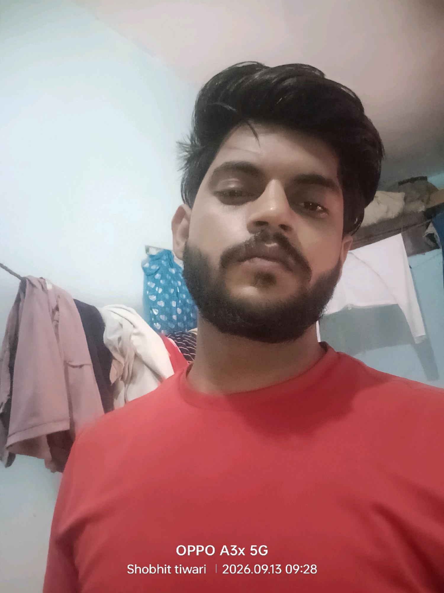 A young South Asian man with a full beard and styled dark hair is pictured from the chest up, looking directly at the camera. He is wearing a plain red t-shirt. The background appears to be an indoor setting, possibly a room, with a light-colored wall. To the left of the man, a rack of clothing is visible, with several garments hanging on it, including what appear to be shirts or jackets in shades of pink and dark blue. Behind the man and to the right, there are some objects on shelves, including a blue patterned cloth or bag and a white sheet or cloth. The image also contains text overlay at the bottom, indicating "OPPO A3x 5G", "Shobhit tiwari", and "2026.09.13 09:28". The location is identified as Kharkhauda, India.