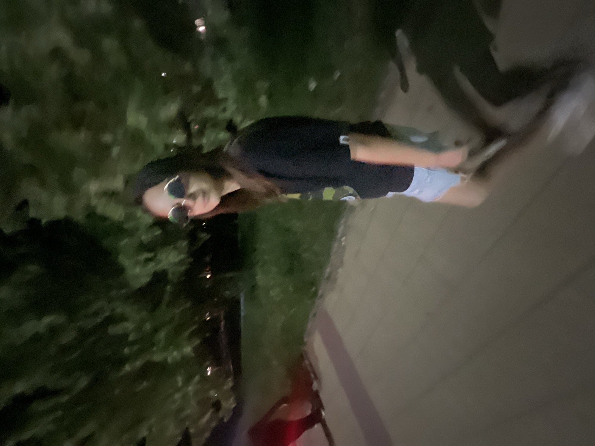This image, taken in the city of Unknown, Unknown, depicts an outdoor scene at night under low light conditions. The primary subject is a female individual, likely in her late teens or early twenties, with long, dark hair, standing or walking on a paved surface. She wears round, reflective sunglasses, a dark short-sleeved top with a light-colored design on the left chest, and light blue denim shorts. Her body is angled slightly to the left, and her head is turned towards the viewer. On the far left, the lower legs of another blurred individual, illuminated by a red light source, are partially visible. The foreground and midground consist of a light-colored paved path, possibly concrete or wood planks, with discernible lines. To the right of the path is a strip of dark grass or low-lying vegetation. The background features dark, blurred foliage, indicative of trees, with faint, distant artificial lights suggesting an urban or suburban environment. The entire image exhibits significant motion blur, particularly in the background and lower portions of the subjects, consistent with movement during a low-light capture.