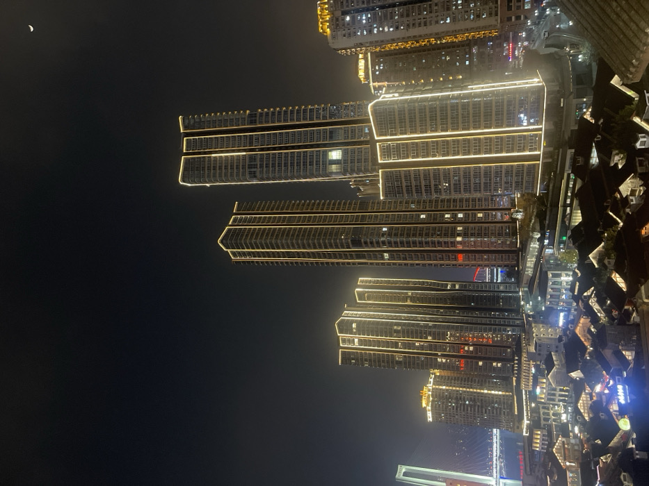 The image presents a nighttime urban landscape located in Yuzhong District, Chongqing, China, observed from an elevated perspective. The upper section of the frame features a dark, clear sky with a slender crescent moon visible in the upper right. The midground and background are populated by a dense array of modern skyscrapers.
Multiple high-rise buildings, primarily located centrally and extending to the right, are prominently illuminated with bright yellow-gold LED strip lights outlining their vertical and horizontal architectural contours. Interior lights are visible through windows on many of these structures. To the far left, a tall, slender white pylon, indicative of a cable-stayed bridge (likely the Dongshuimen Bridge or Qianximen Bridge), rises above a body of water in the distance.
In the foreground, situated below the towering skyscrapers, is a cluster of smaller, multi-story buildings constructed in a traditional Chinese architectural style, characterized by dark, sloped roofs and intricate facades. These lower structures are also lit, displaying various residential and commercial lights, including at least one blue neon sign with Chinese characters visible on a building in the lower left. No human figures or discernible actions are present within the scene.