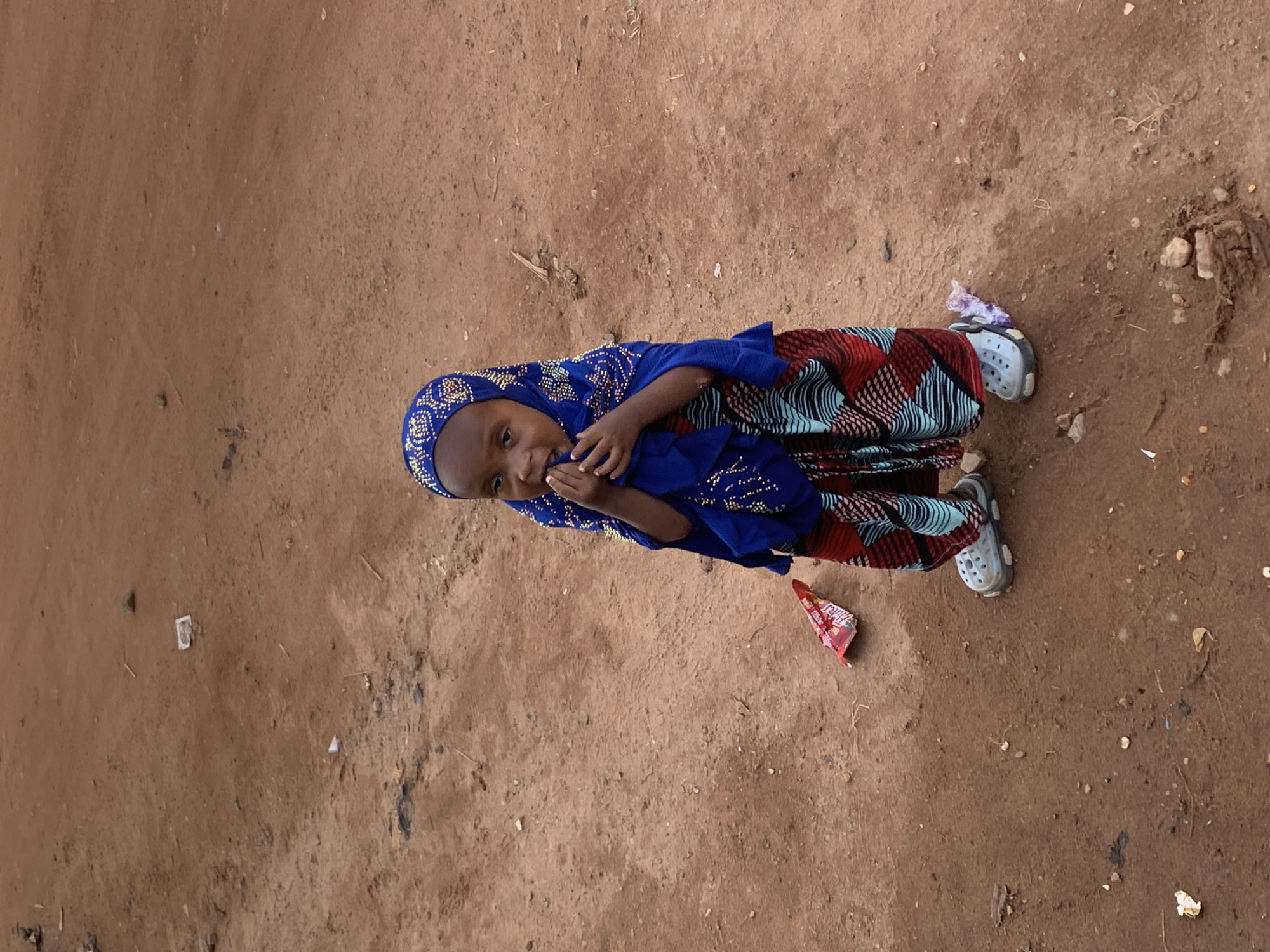 A young child, likely a girl, is standing in an outdoor environment with a dirt ground. She is wearing a blue headscarf adorned with decorative white and gold patterns, and a colorful patterned skirt with red, blue, and black designs. On her feet, she wears light blue Croc-style shoes. The child has her hands near her mouth. The ground is composed of reddish-brown dirt with scattered small rocks and debris, including a red and white wrapper to the child's left and a small plastic bag near her right foot. The background is out of focus and appears to be a continuation of the dirt terrain. The lighting suggests it is daytime. The scene is likely in an unpaved area of Abuja, Nigeria, characterized by its earth-toned soil and sparse vegetation or debris.