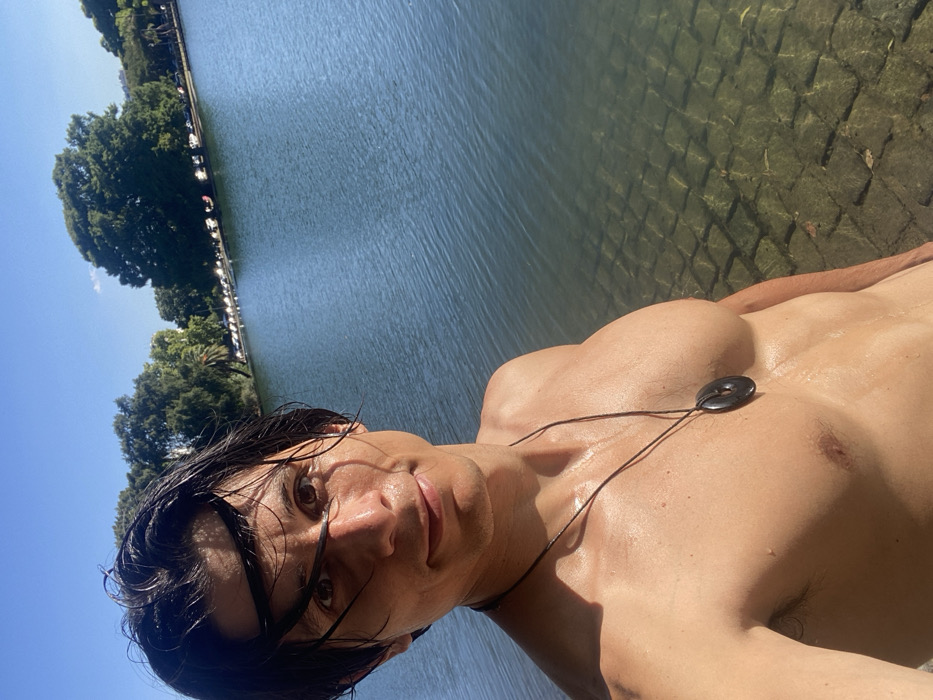 A shirtless man, glistening with sweat, is positioned in front of a body of water. The water reflects the sky, revealing submerged stone or tiled paving along the edge. Lush green trees and foliage line the far side of the water. The man wears a dark necklace with a circular pendant. The image is taken at an oblique angle, capturing a close-up of the man's face and upper torso. The lighting is bright and sunny, highlighting the texture of the water and the sheen on the man's skin.