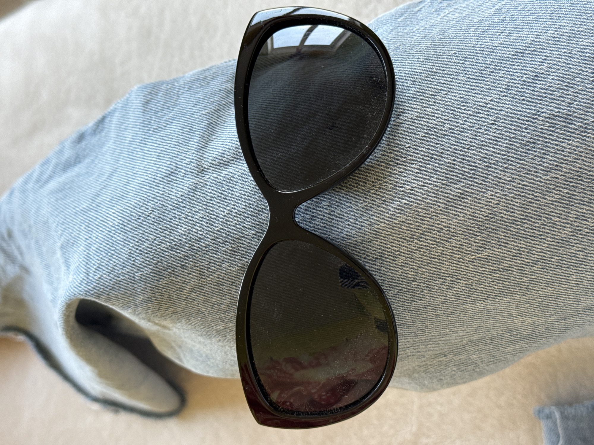 A pair of black, cat-eye style sunglasses rests diagonally on a light blue denim fabric, likely part of a pair of jeans or a jacket. The denim occupies the majority of the frame, displaying its characteristic weave pattern with horizontal white and blue threads. The sunglasses are centered on the denim, with their frames appearing smooth and shiny, though the lenses show some dust or smudges.
In the reflection of the top lens, a bright, rectangular light source, possibly a window or skylight, with a visible grid pattern is apparent, indicating an indoor setting during daylight hours. The bottom lens also shows reflections, though less distinctly, appearing to be of a light-colored, possibly textured surface like a ceiling or wall, and hints of a darker, perhaps reddish object or reflection.
The background around the denim is a soft, out-of-focus beige or light cream color, suggesting a surface such as a couch, bed, or floor. To the bottom left, an open cuff or folded edge of the denim is visible, showing the lighter underside of the fabric. No people are visible, nor is any specific activity taking place other than the sunglasses being set down. There is no visible text. The overall scene is a close-up, focusing on the textures of the objects and the subtle reflections.