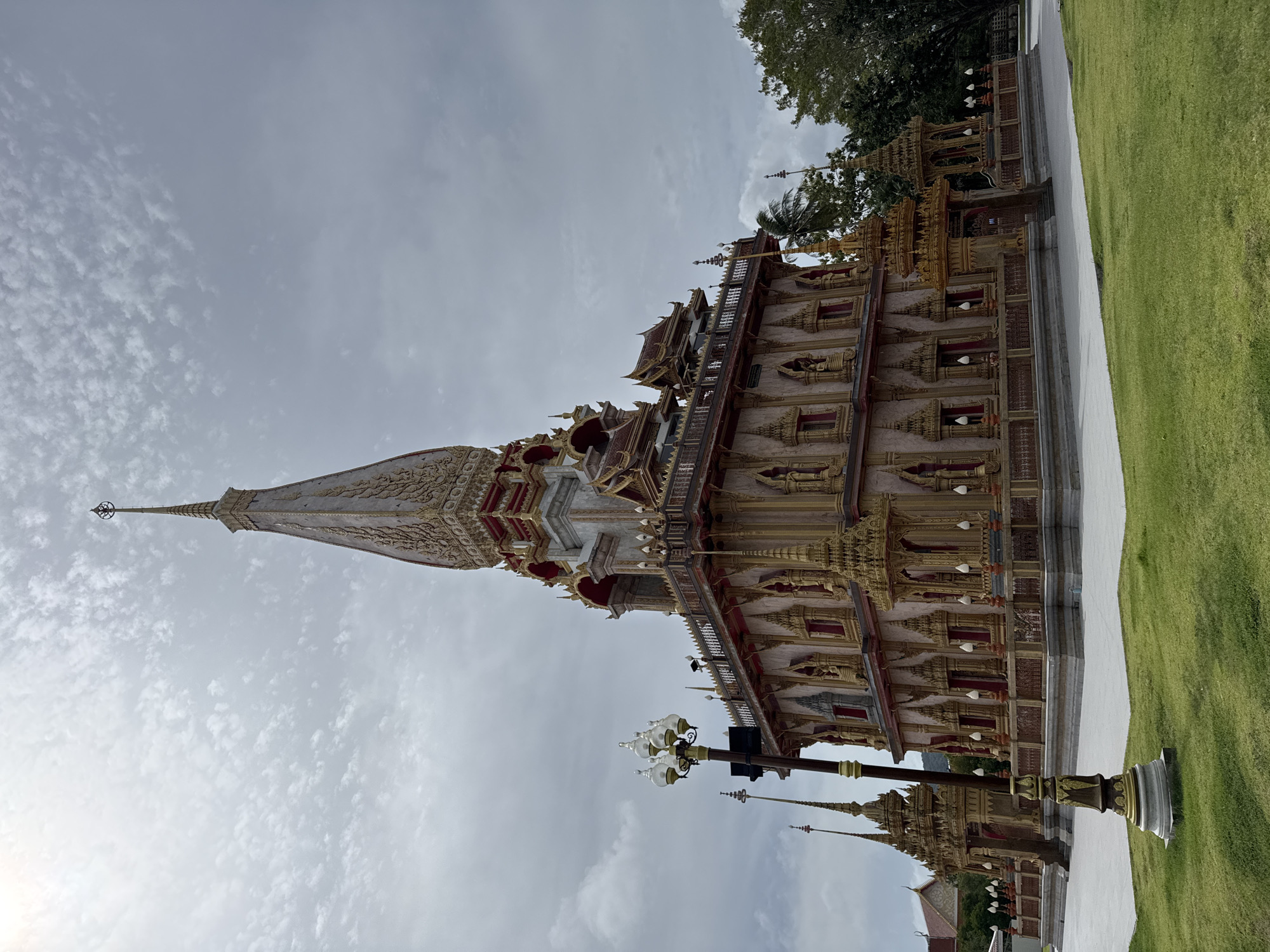 This is a low-angle, exterior, full shot of the Chalong Temple (Wat Chalong) in Chalong, Thailand. The ornate, multi-tiered temple structure dominates the frame, featuring intricate gold detailing, red accents, and a tall, pointed spire reaching towards a cloudy, overcast sky. The temple's facade is adorned with numerous statues and decorative elements. In the foreground, a well-maintained green lawn stretches across the scene, with a vintage-style lamppost situated to the left of the temple. To the right of the temple, lush green trees are visible. The overall impression is one of grandeur and cultural significance.