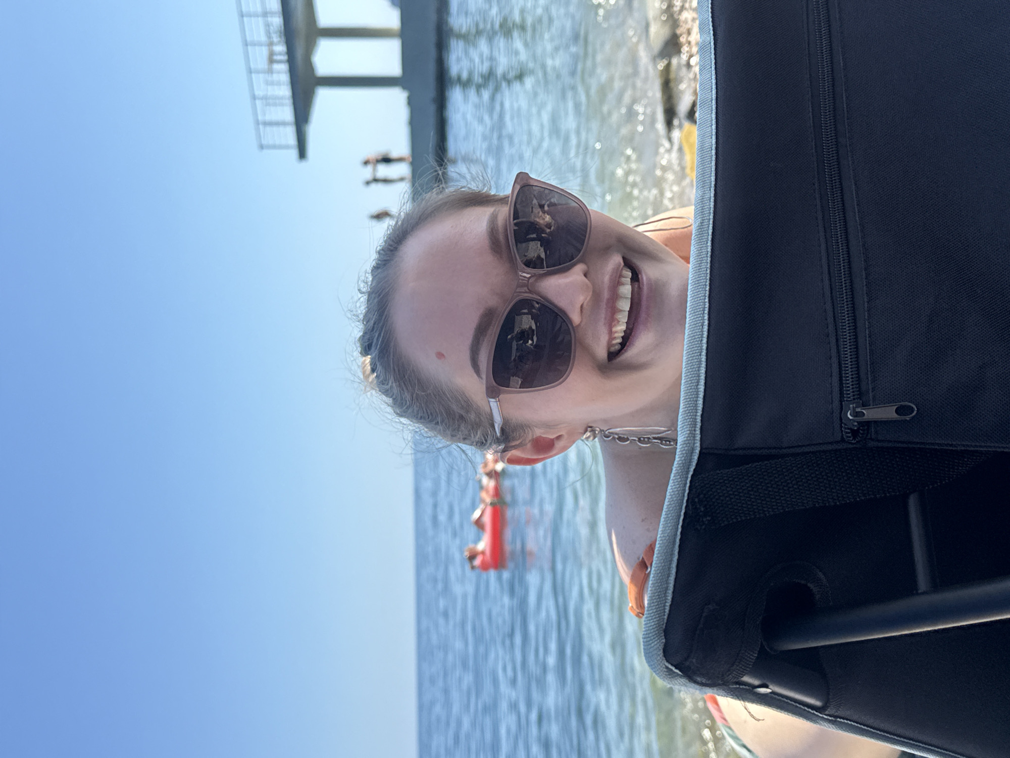 The image shows a woman sitting on a beach in Utios, Ukraine. She is wearing sunglasses and a swimsuit, and she is smiling at the camera. The beach is rocky, and the water is clear. In the background, there is a diving platform and several people are visible on it. The sky is blue and clear. The woman appears to be happy and relaxed.