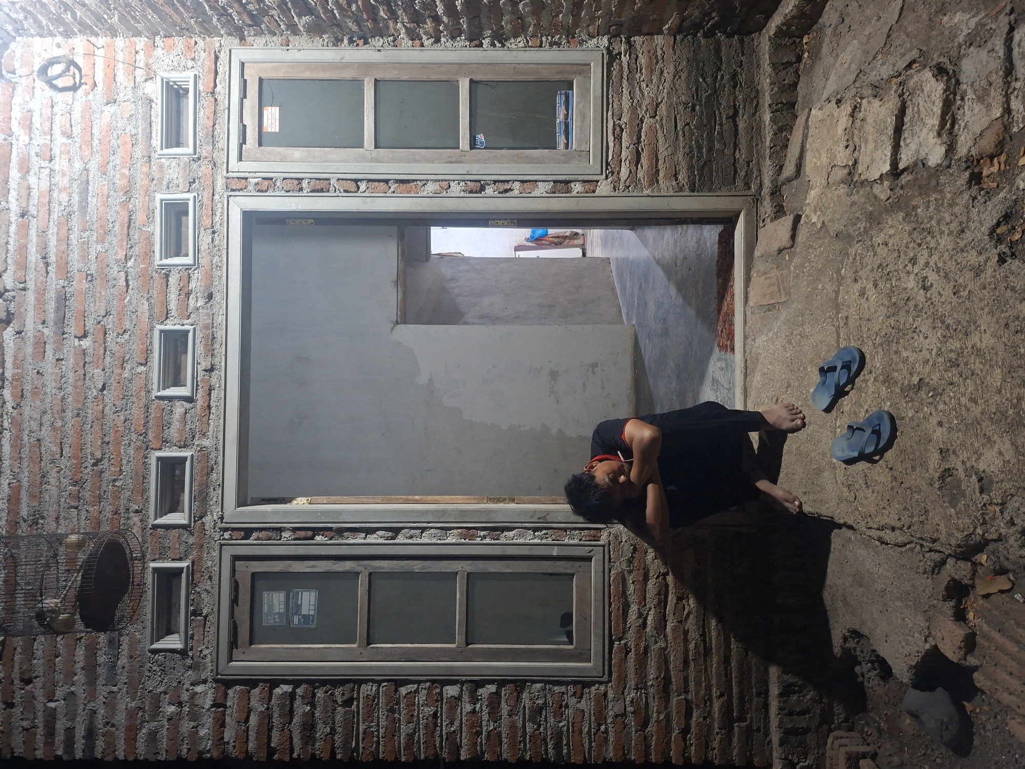 A person wearing a black t-shirt with a red collar is sitting on the ground outside a brick building with their head down, elbows on their knees and hands clasped. The person is barefoot. Two pairs of blue flip-flops are on the ground near them. The building has two windows and an open doorway. A bird cage hangs on the wall above one of the windows. The ground is made of concrete and brick. The scene appears to be in the city of Tongaswetan, Indonesia.