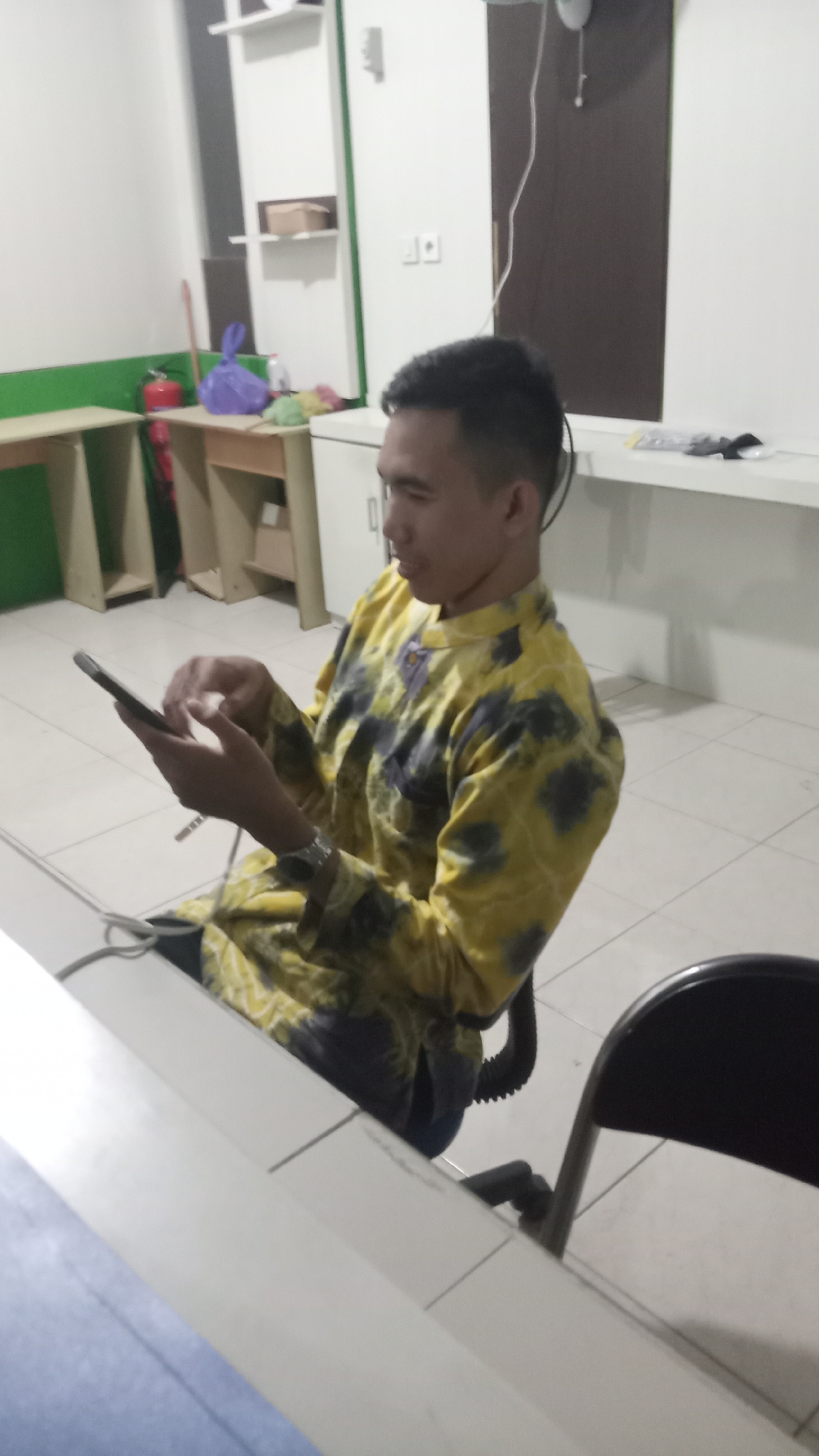 A young man is seated in a black chair, facing left, and using a smartphone. He is wearing a yellow and dark patterned long-sleeved shirt. The man is in an indoor environment with tiled flooring and white walls. In the background, there are light-colored shelves with various items, including a red fire extinguisher, a blue plastic bag, and other unidentifiable objects. A white desk is visible in the foreground. The lighting appears to be artificial, casting soft shadows. The scene is consistent with an office or a common area within a building in Keraton, Indonesia.