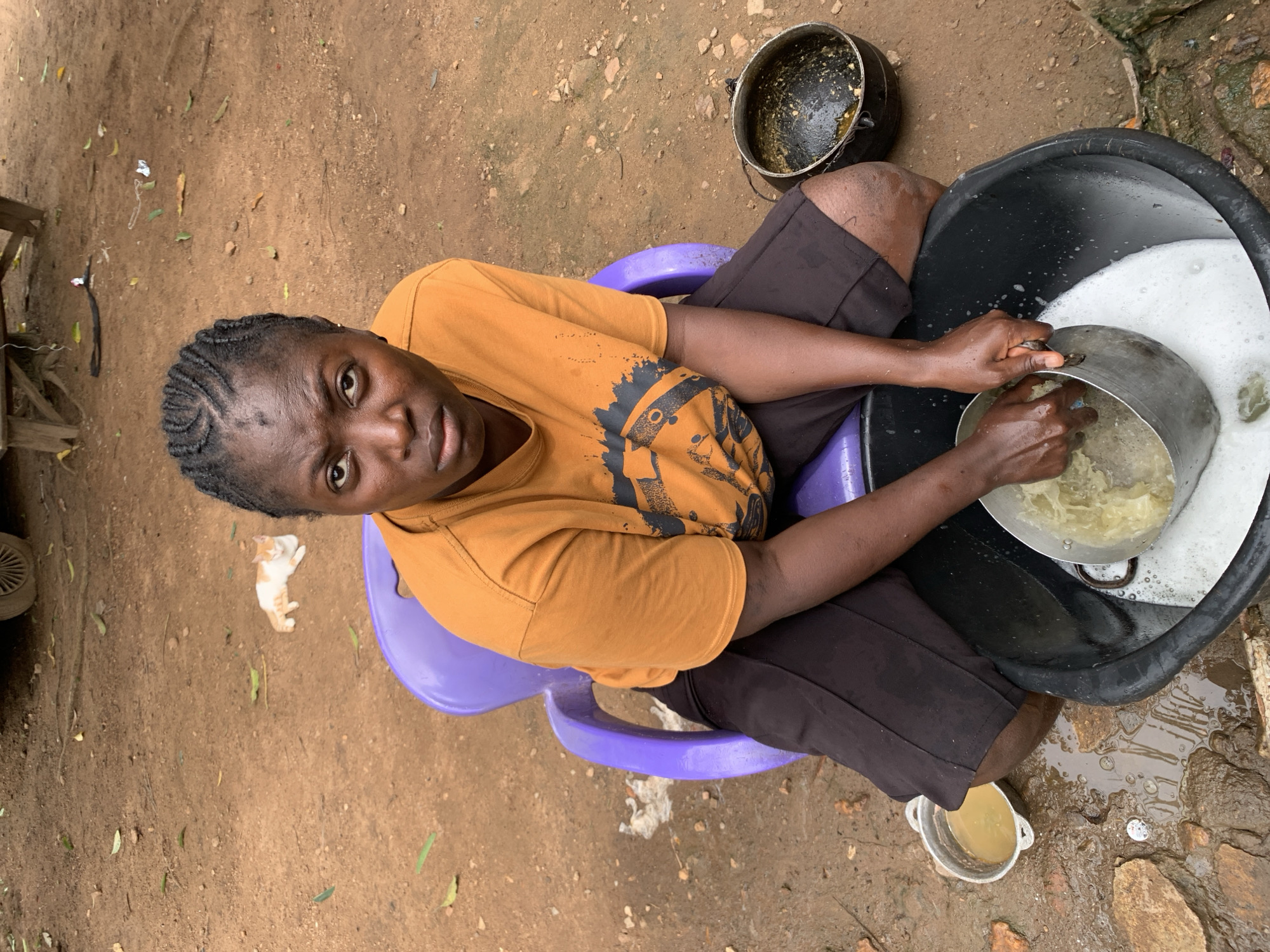 A woman with braided hair sits on a purple plastic chair outdoors in Akwanga, Nigeria, washing dishes in a large black basin filled with soapy water. She wears an orange t-shirt with a graphic design and black shorts. In her hands, she holds a metal pot, scrubbing it. To her left, a smaller metal bowl contains a yellowish liquid. To her right, another dark pot sits on the ground. The ground is reddish-brown dirt with scattered stones and some fallen leaves. In the background, a white and orange cat lies on the dirt, and a portion of a vehicle wheel is visible.