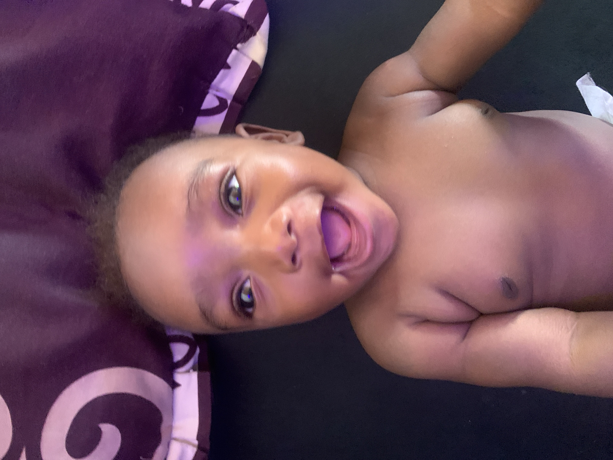 A baby with brown skin is lying on its back, facing the camera. The baby has its mouth open wide, appearing to be smiling or laughing. A dark purple pillow with a white swirl design is visible on the left side of the image. The background is a dark surface. There is a white tissue or piece of cloth on the baby's lower right side. The location is unknown.