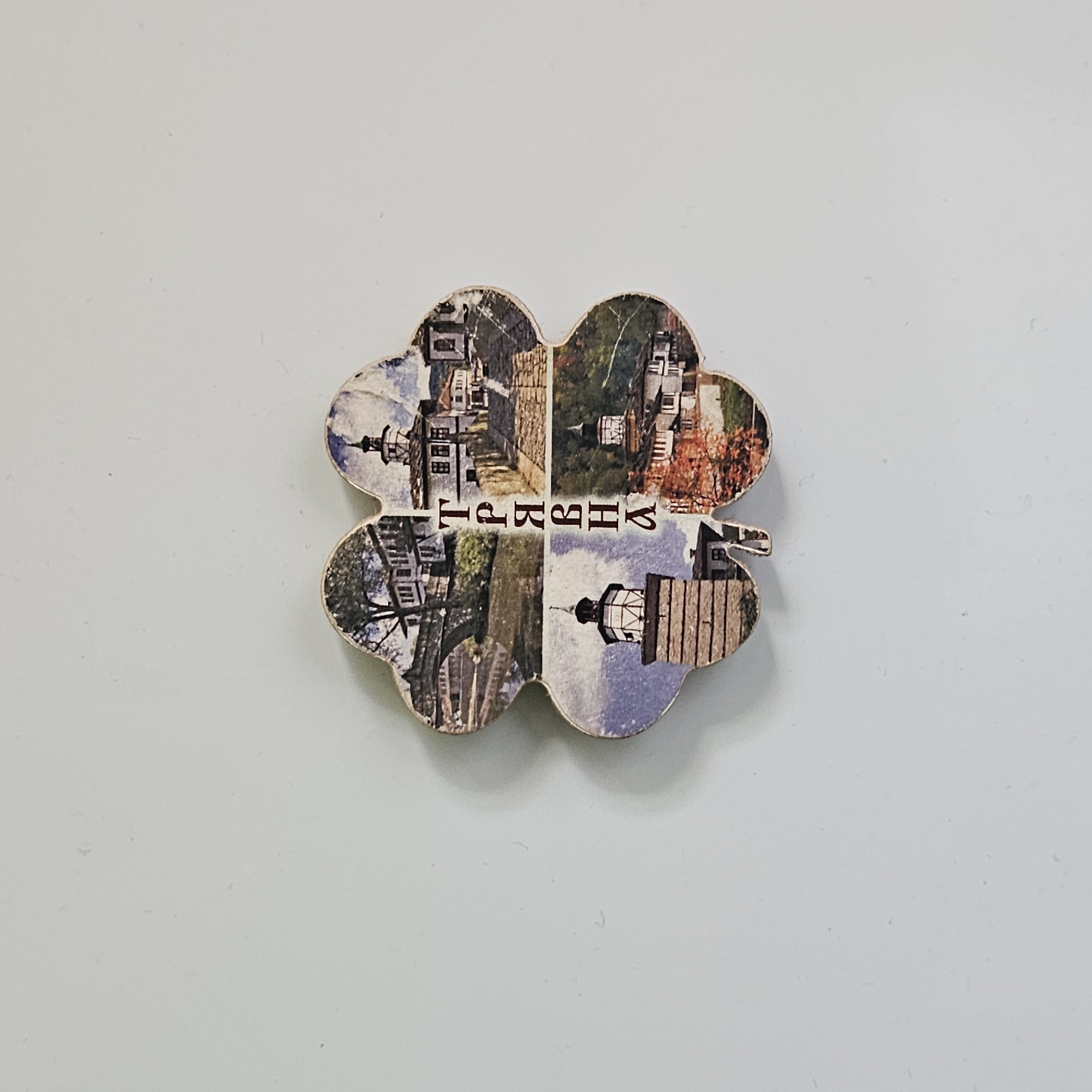 A four-leaf clover-shaped souvenir magnet displays the name "Stara Zagora," a city in Bulgaria. The clover is divided into four sections, each containing a different photograph. The upper left section shows a building with a tower. The upper right section features buildings and autumn foliage. The lower left section shows buildings and trees. The lower right section shows a building with a tower. The magnet is on a light-colored surface.