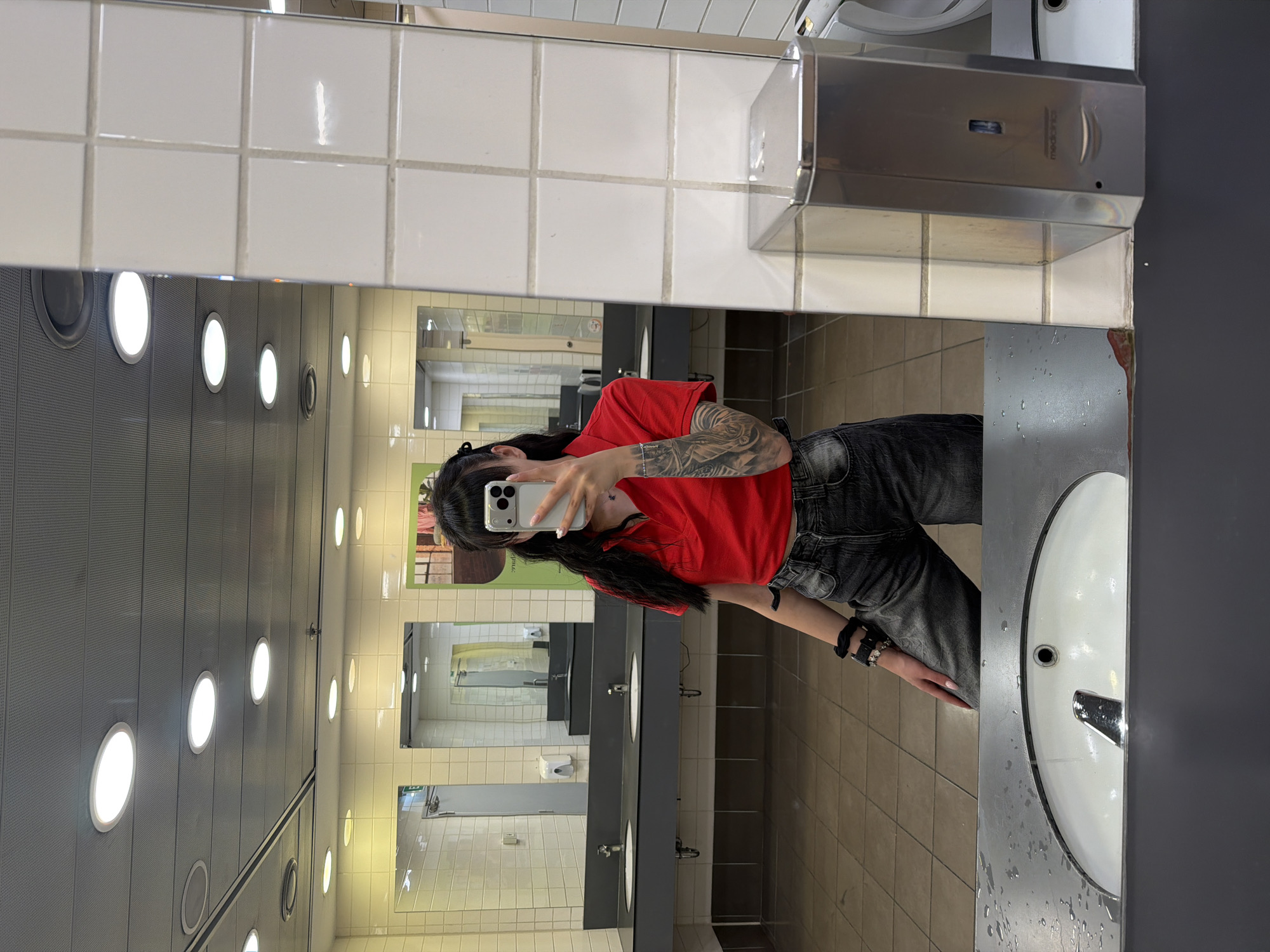 The image shows a young woman with long dark hair, wearing a red polo shirt and grey jeans, taking a selfie in a public restroom. She has tattoos on her arm. The restroom has white tiled walls, a tiled floor, and multiple sinks. The woman is standing in front of a mirror, holding a smartphone. The environment appears to be a public facility, possibly a shopping mall or a train station. There is no visual information in the image to suggest the location is in Plovdiv, Bulgaria.