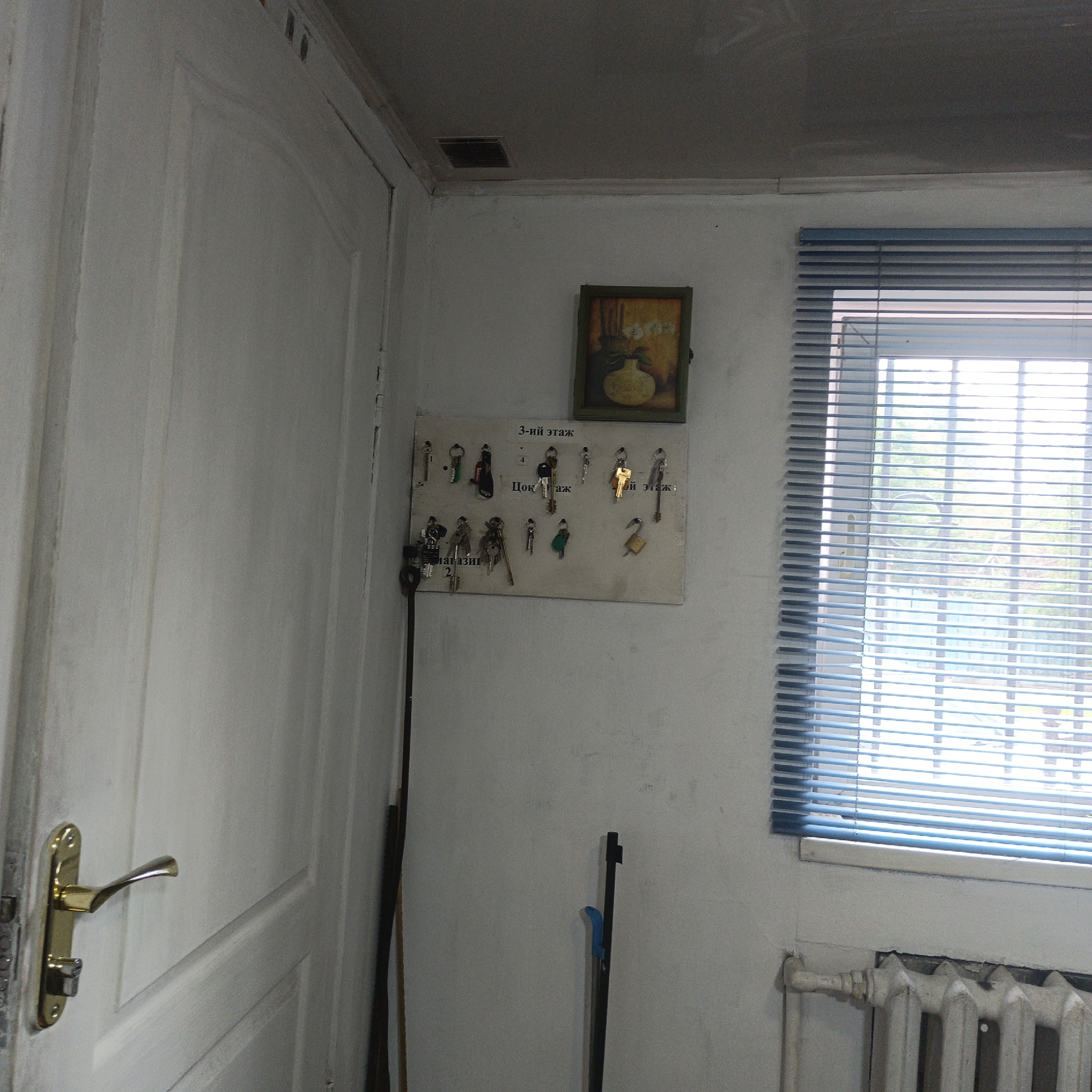 The image depicts an interior scene, possibly a hallway or utility area, with elements suggestive of a residential or communal building in Ulan-Ude, Russia. On the left, a white door with a brass handle and lock mechanism is visible. The wall to the right of the door is painted white and has a small framed picture of a vase with flowers hanging on it. Below the picture is a key rack holding numerous keys, some of which are labeled with Cyrillic text. The labels appear to indicate floor numbers and room designations such as "3-ий этаж" (3rd floor), "Цок. этаж" (basement floor), and "магазин" (shop). A long, thin dark object, possibly a mop handle or a broom, leans against the wall near the key rack. Further to the right, a window covered with horizontal blue blinds is present. Outside the window, trees and some structures can be faintly seen. Below the window is a white cast-iron radiator. The ceiling appears to be a dark, textured material with a ventilation grate in the corner. The overall impression is one of a functional, slightly dated interior space.