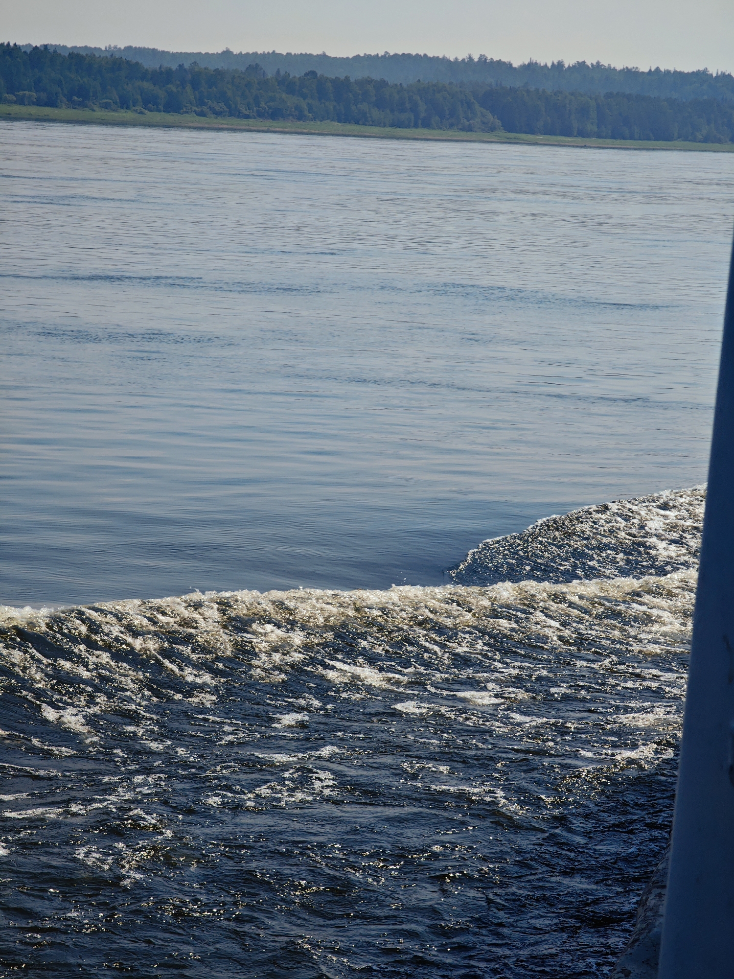 A high-angle, medium shot captures the wake of a vessel moving through a wide, calm body of water, likely a river, with a dense forest bordering the far bank. The water in the foreground is dark blue and choppy, filled with white foam and bubbles created by the boat's movement, reflecting the sunlight. Further out, the water transitions to a lighter, serene blue with subtle ripples. The distant shore is lined with a thick, dark green forest that stretches across the horizon, meeting a pale, hazy sky. A portion of a grey structure, possibly part of the vessel, is visible on the right side of the frame, extending from the bottom edge upwards and partially obscuring the water. The scene appears to be set in Pogodaevo, Russia, given the context. No people or specific animals are visible. The overall atmosphere is peaceful and natural.